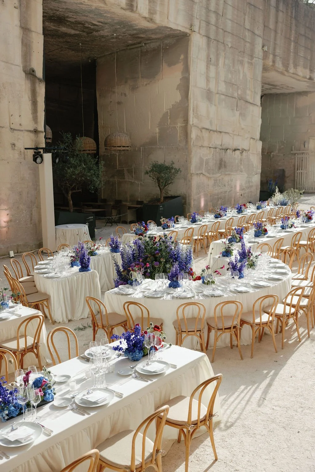Indoor event space with long banquet tables decorated with purple and white floral centerpieces, set with white plates, glasses, and silverware. The space has a high stone or concrete wall and hanging wicker lamps, with natural light.