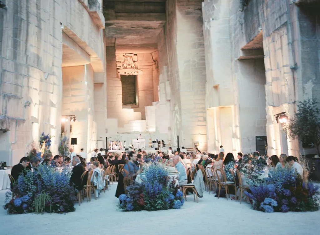 A large indoor venue with high stone walls hosting a formal dinner event with many people seated at round tables decorated with purple and blue floral arrangements.