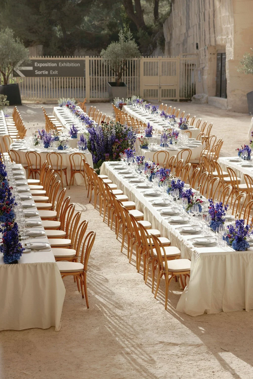 Long banquet tables set with white tablecloths, decorated with blue, purple, and white floral centerpieces, surrounded by wooden chairs, outdoors in a sandy area with a large floral arrangement at the center.