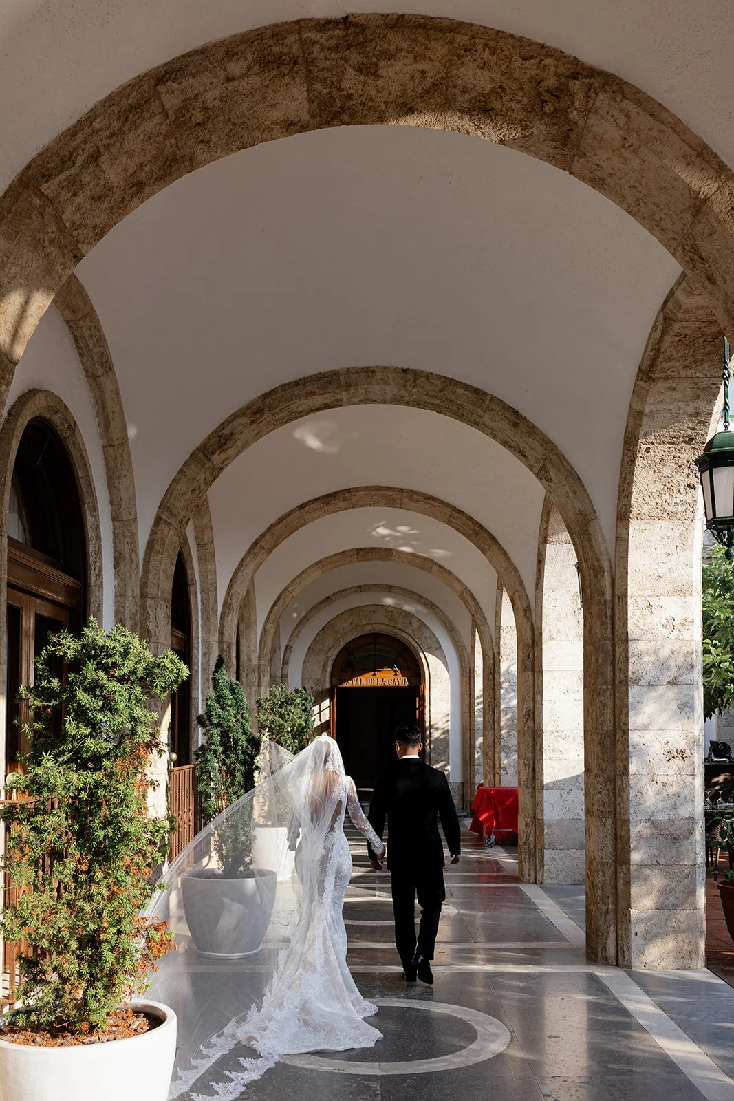 A bride and groom walking hand in hand in an arched outdoor corridor of a stone building, wedding attire, bride in a white lace dress and veil, groom in a black suit, potted plants along the corridor, sunlight and shadows.