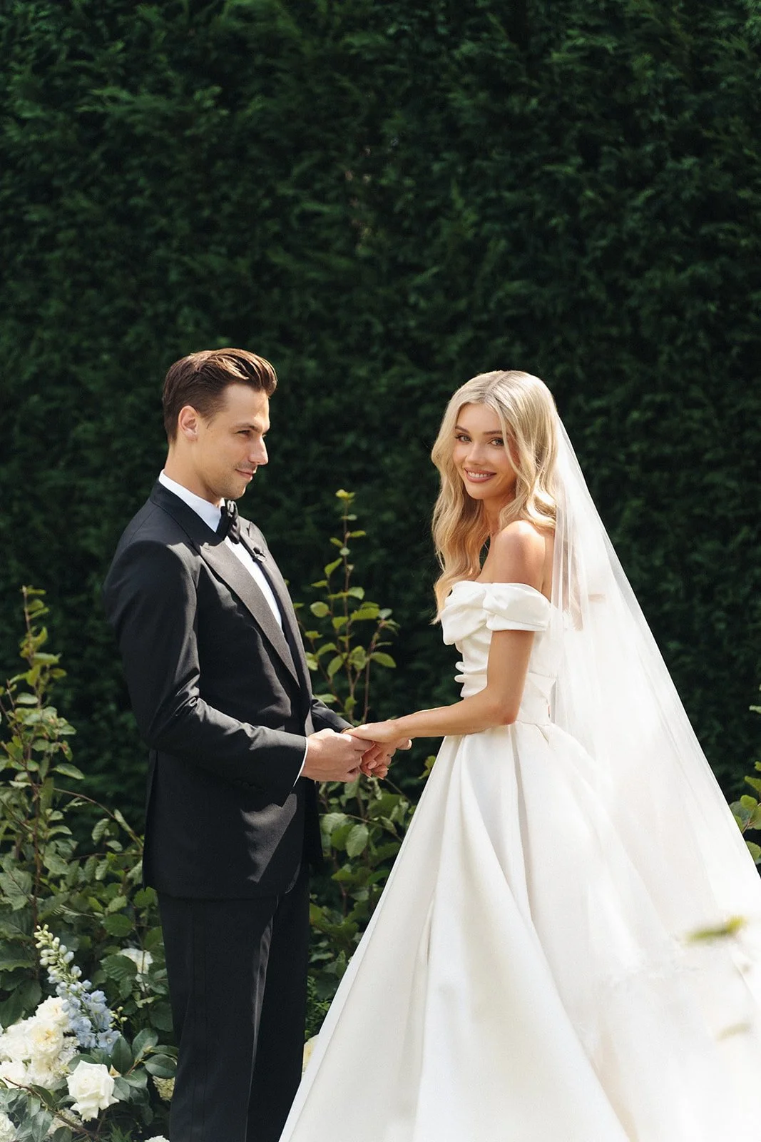 A bride and groom holding hands during their outdoor wedding ceremony, surrounded by greenery.
