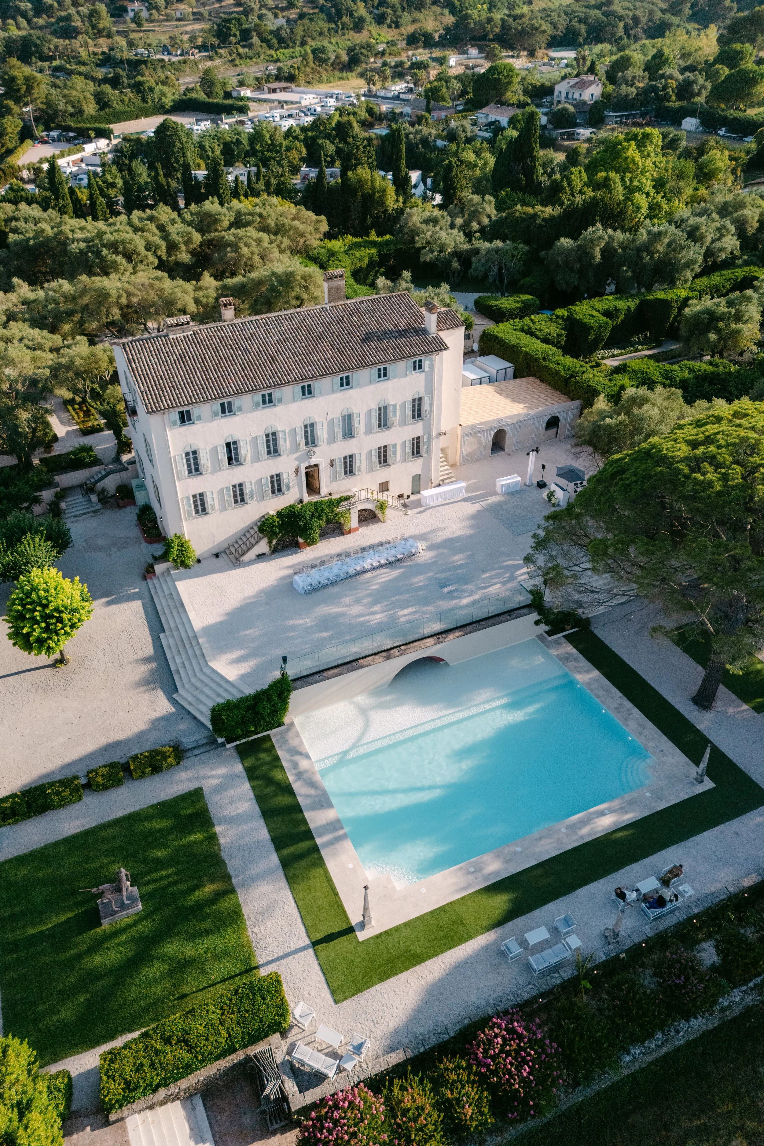 Aerial view of a white building with a tiled roof, a swimming pool, and well-maintained gardens surrounded by trees and greenery.
