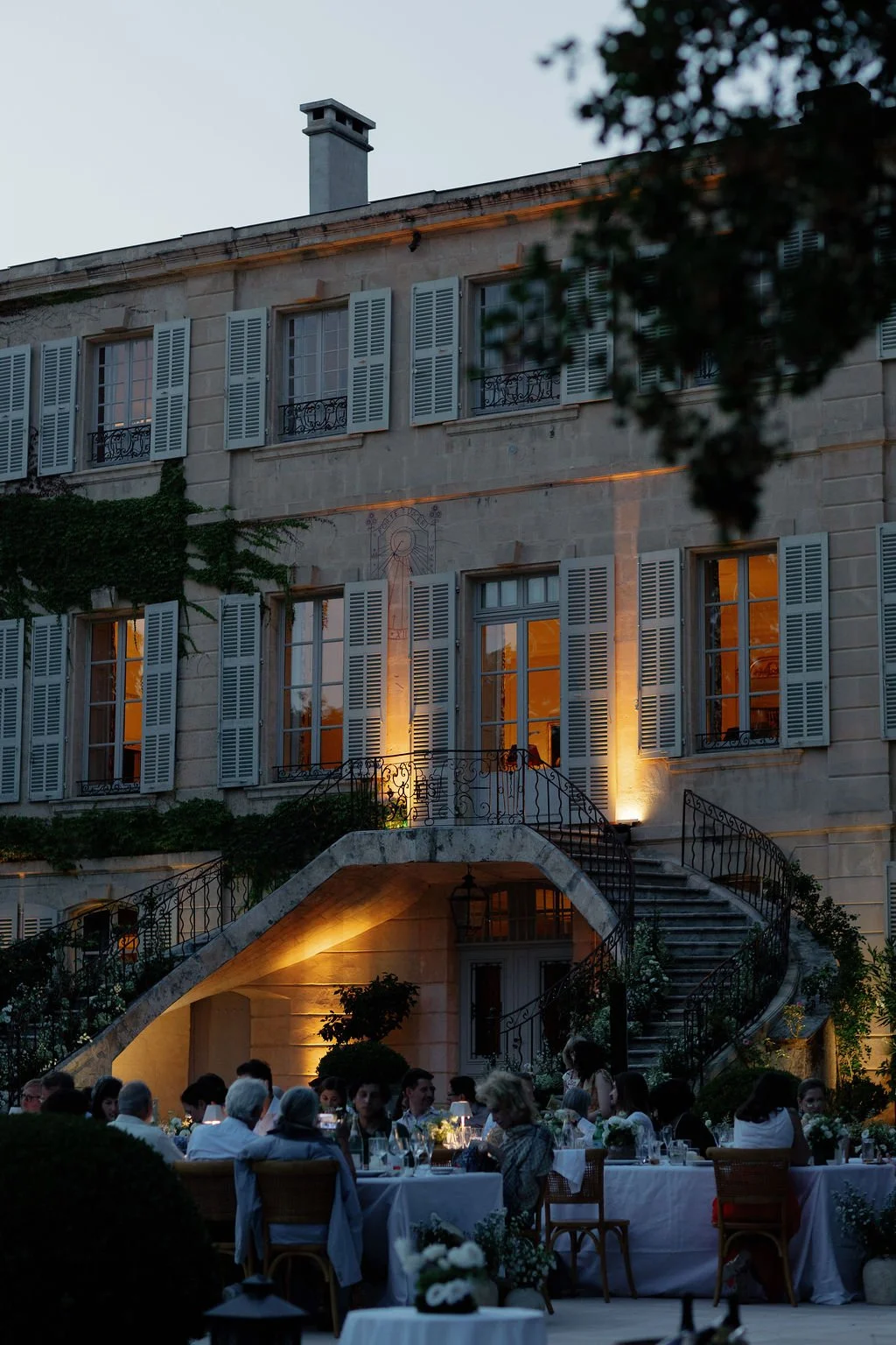 An outdoor dinner event at dusk in front of a large, elegant building with white shutters and wrought iron balconies. The building is illuminated with soft lighting, and tables with people dining are set up on the patio.