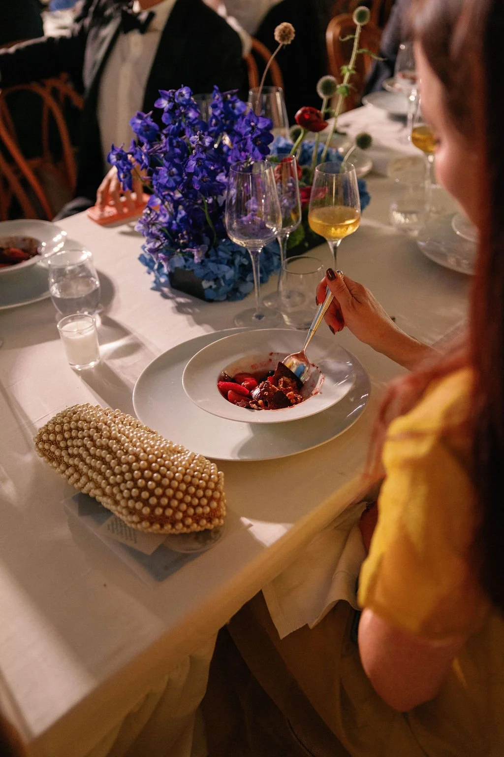 A woman at a dinner table with a white tablecloth, blue and purple floral centerpiece, and wine glasses. She is eating dessert from a white bowl with a spoon. Pearls are on the table, and other guests are visible in the background.