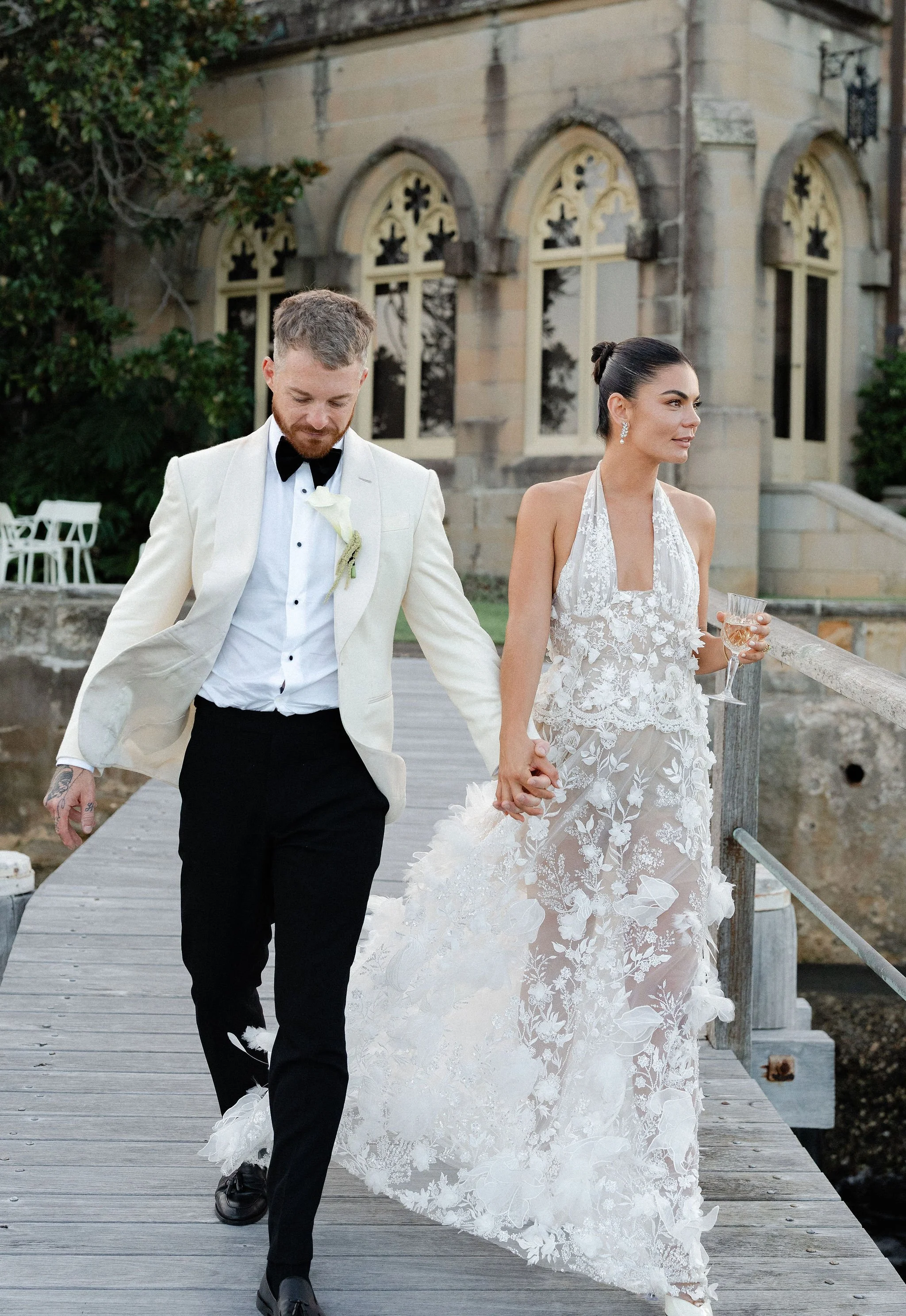 A bride and groom walk hand in hand on a wooden dock, holding a glass of wine, with a historic building with arched windows in the background.