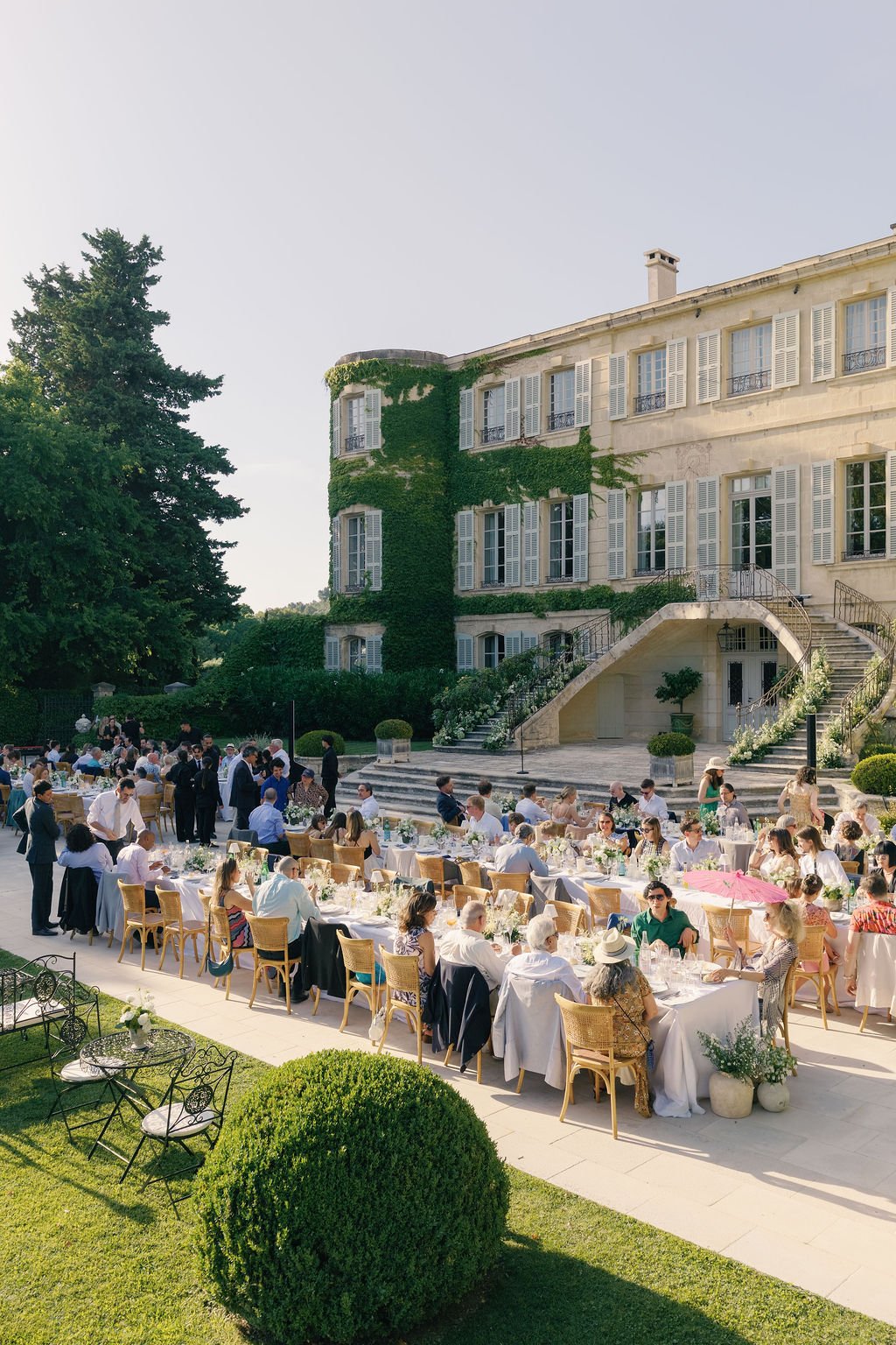 Outdoor wedding reception or banquet with long tables, chairs, and guests in front of a large historic building covered in green ivy, with a staircase decorated with flowers and greenery.