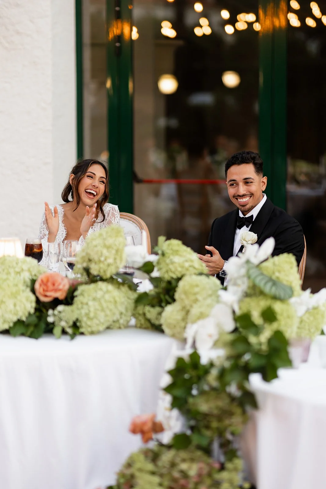 A joyful bride and groom sit at a wedding reception table decorated with white and green floral arrangements, laughing and smiling.