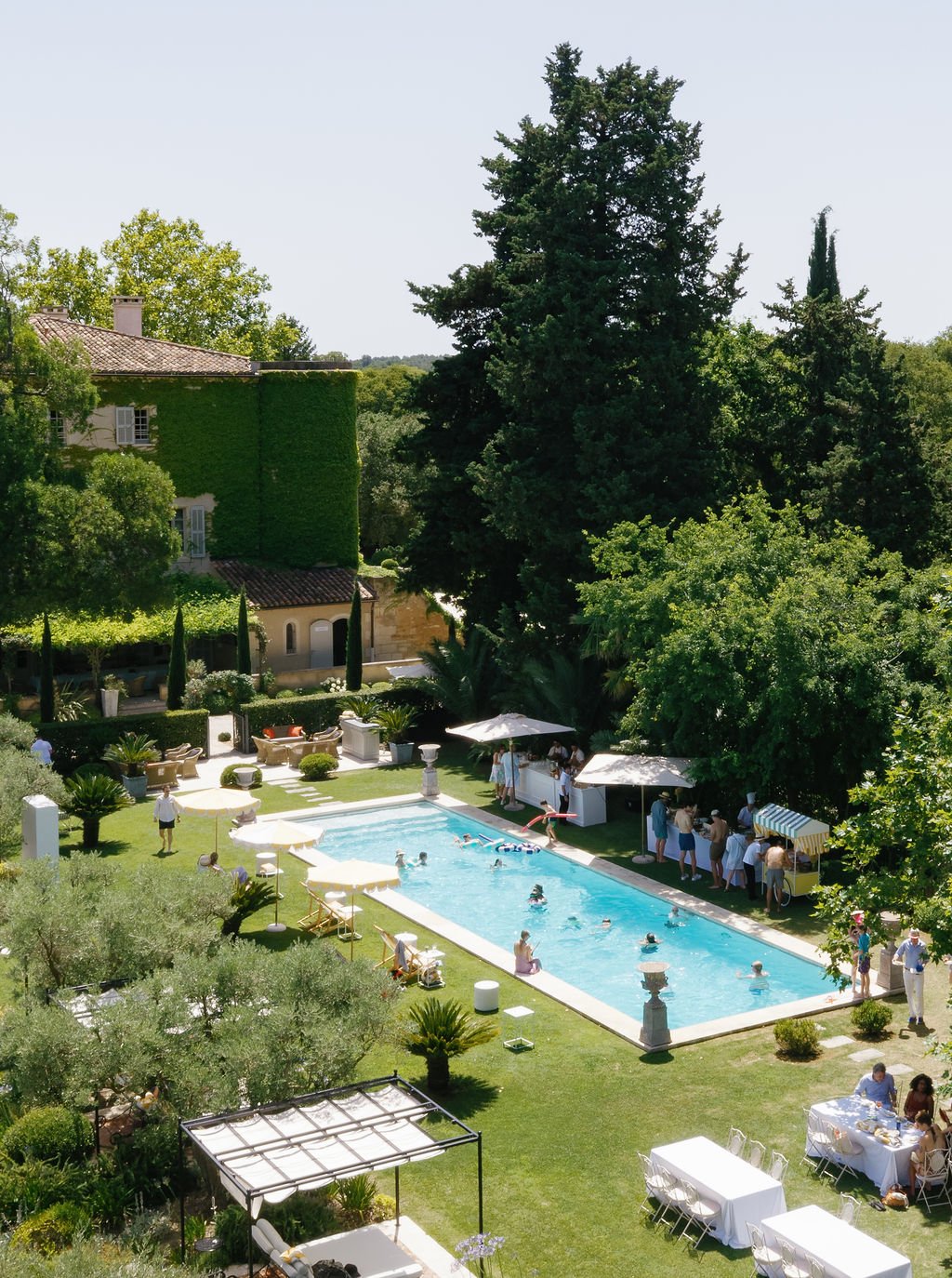People swimming and socializing at a sunny outdoor pool party in a lush green garden, with trees and a house in the background.