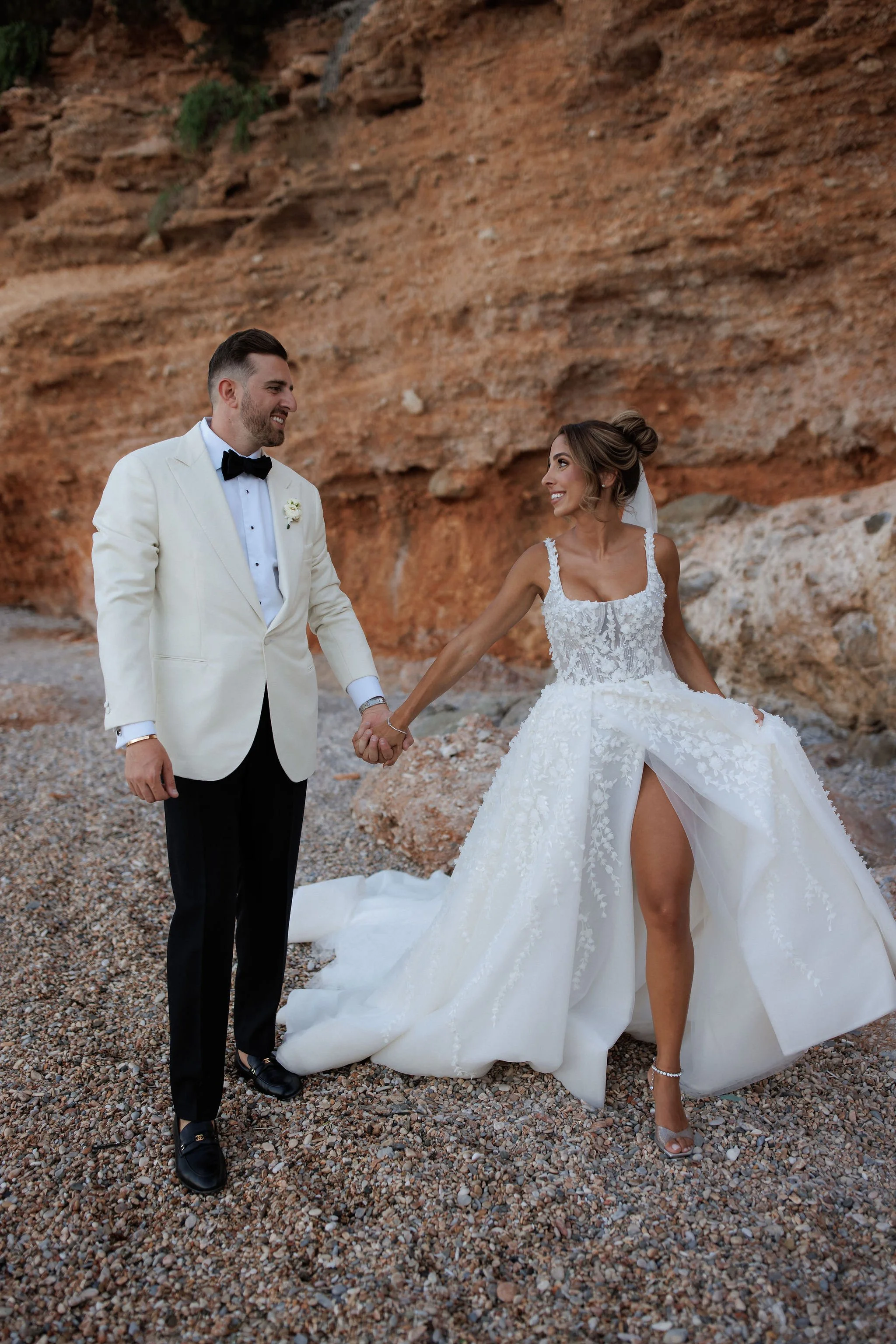 A happy bride and groom holding hands on a pebble beach with a rocky cliff behind them, the bride wearing a white wedding gown with a high slit and the groom in a white tuxedo jacket with black pants.