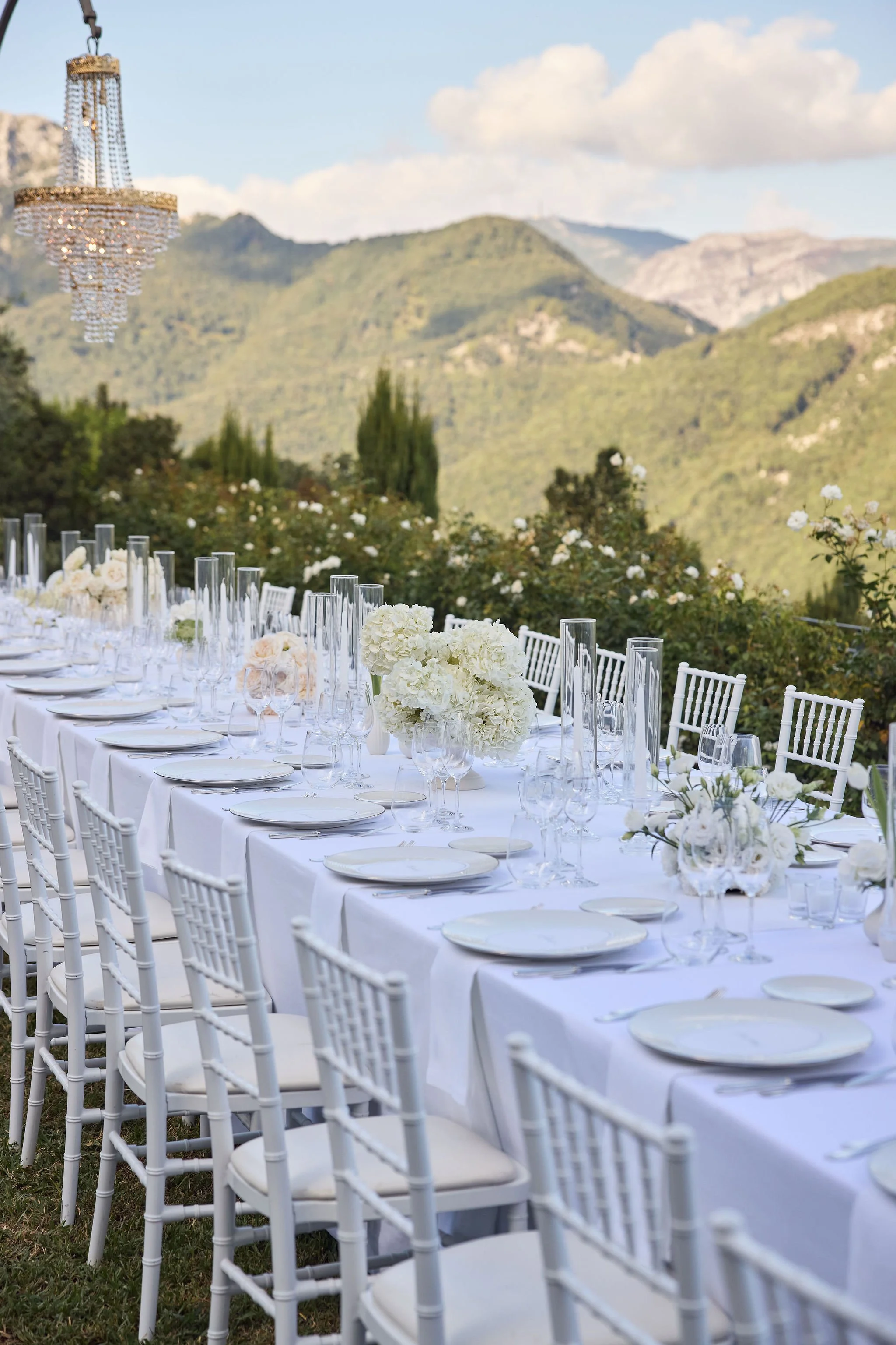 Wedding reception table set outdoors with white tablecloth, white chairs, floral centerpieces, glassware, and plates with a mountainous landscape in the background.