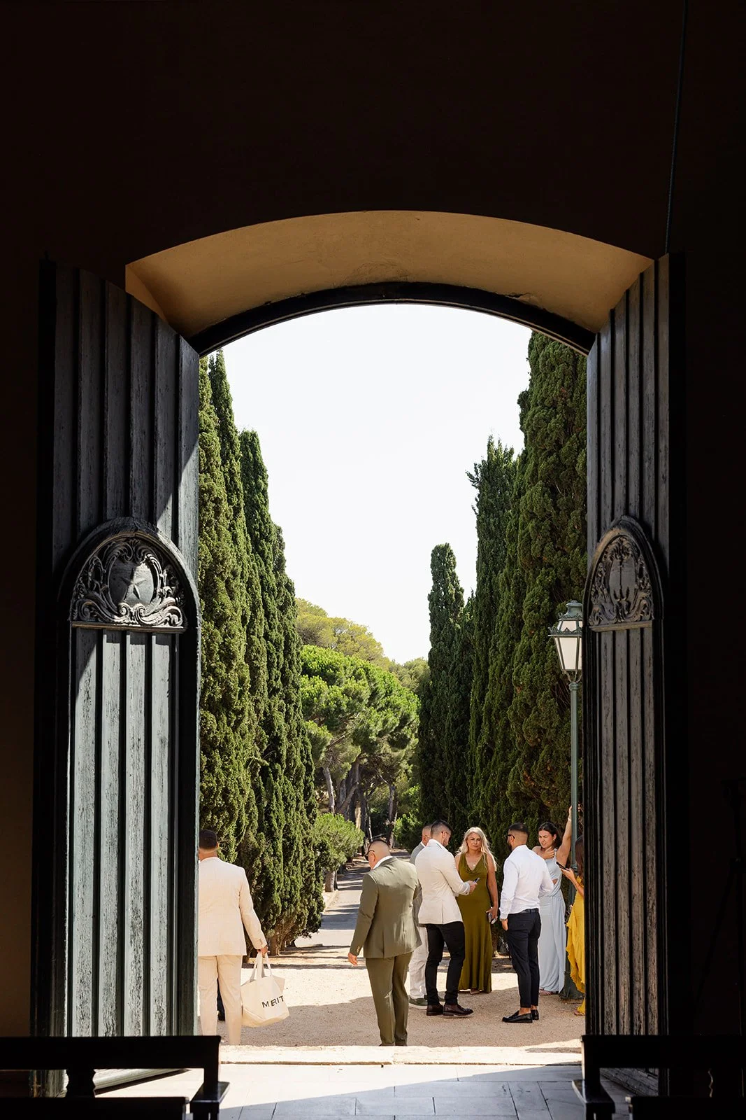 Open large wooden gates reveal a formal outdoor gathering with people dressed in suits and dresses, surrounded by tall, green cypress trees under a bright sky.