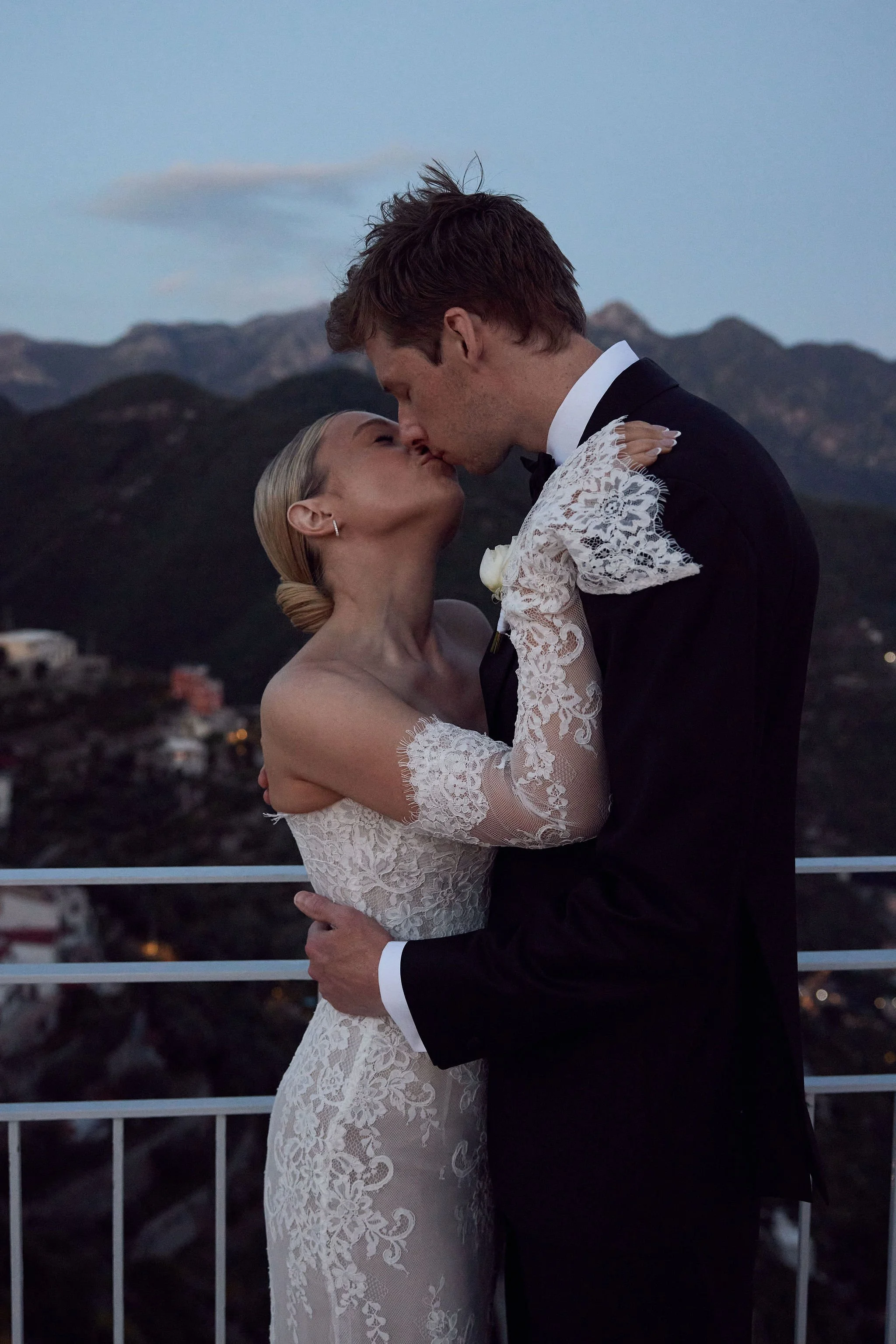 A bride and groom sharing a kiss on a balcony at sunset with mountain scenery in the background.