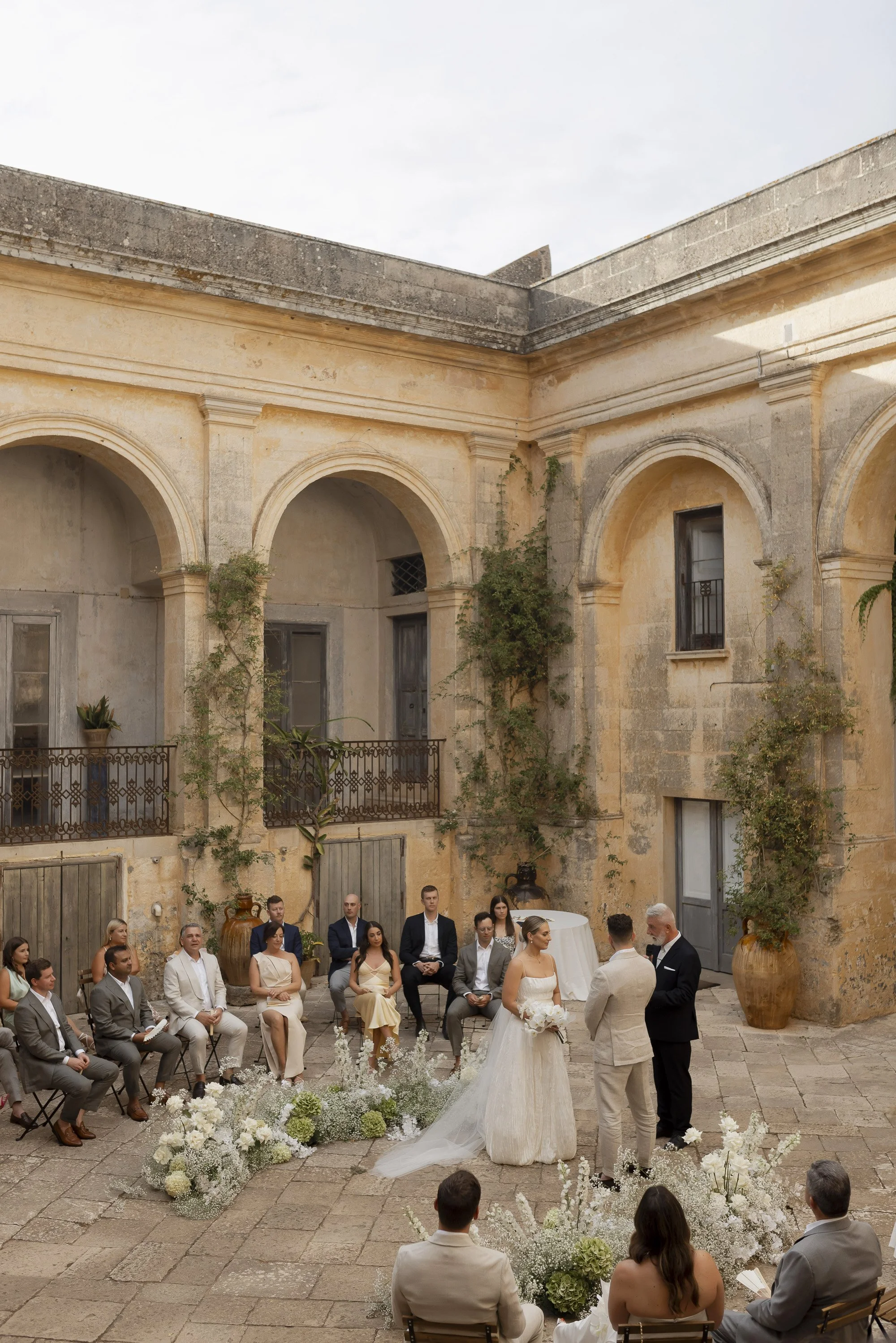 A wedding ceremony taking place outdoors in a historic courtyard with stone walls, arches, and potted plants, featuring a bride, groom, officiant, and guests.