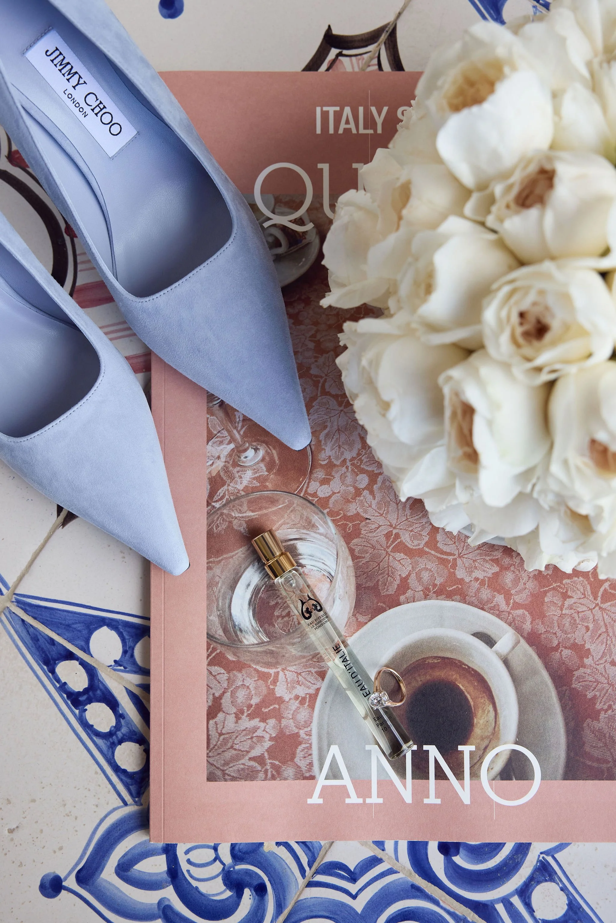 Blue high heels, a bouquet of white roses, a glass bottle, a small cup of espresso on a table with a floral tablecloth, and a magazine or poster with the word 'Italy' on it.
