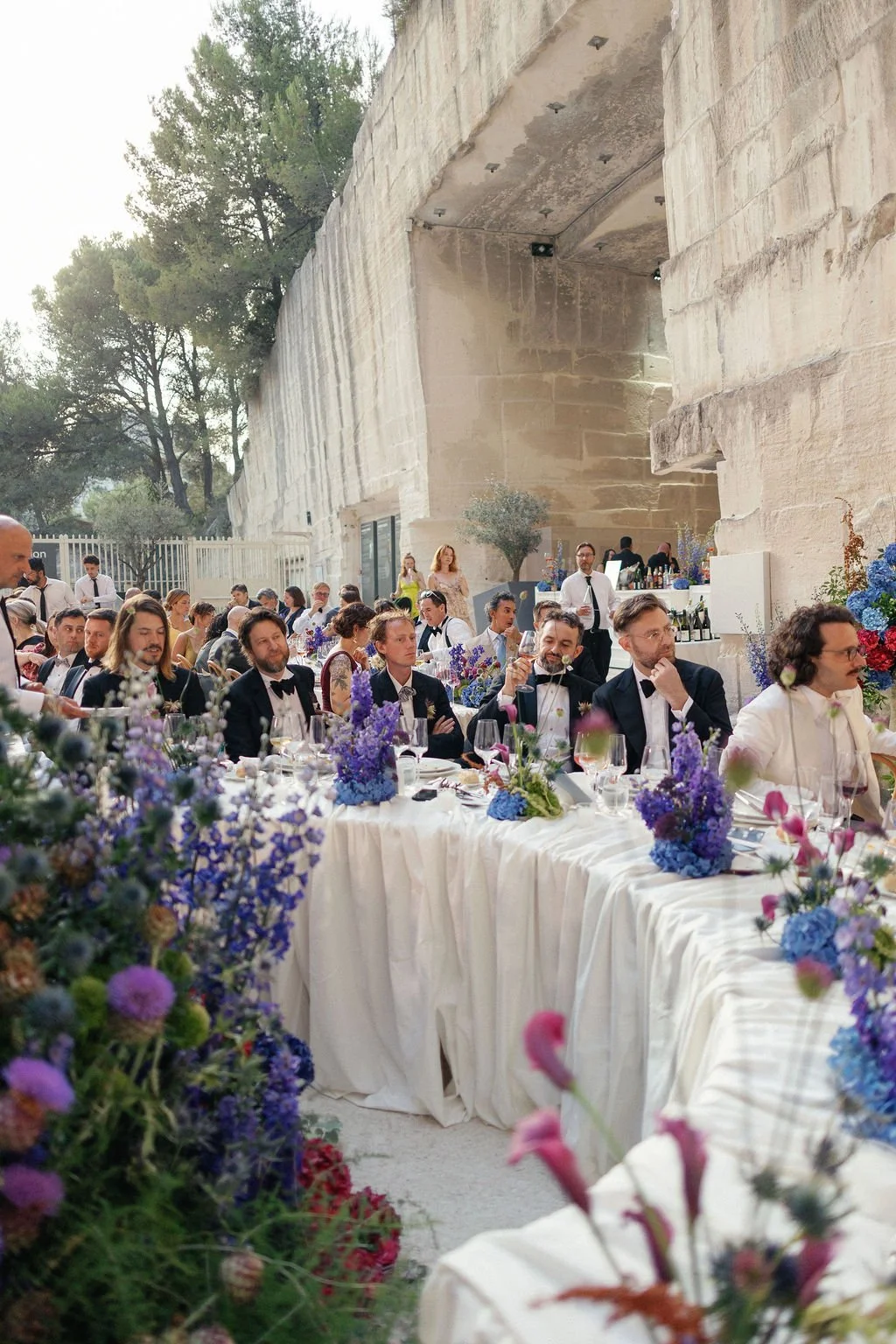 People attending a formal event or wedding reception with floral decorations, seated at decorated tables in an outdoor setting with large stone walls and trees in the background.