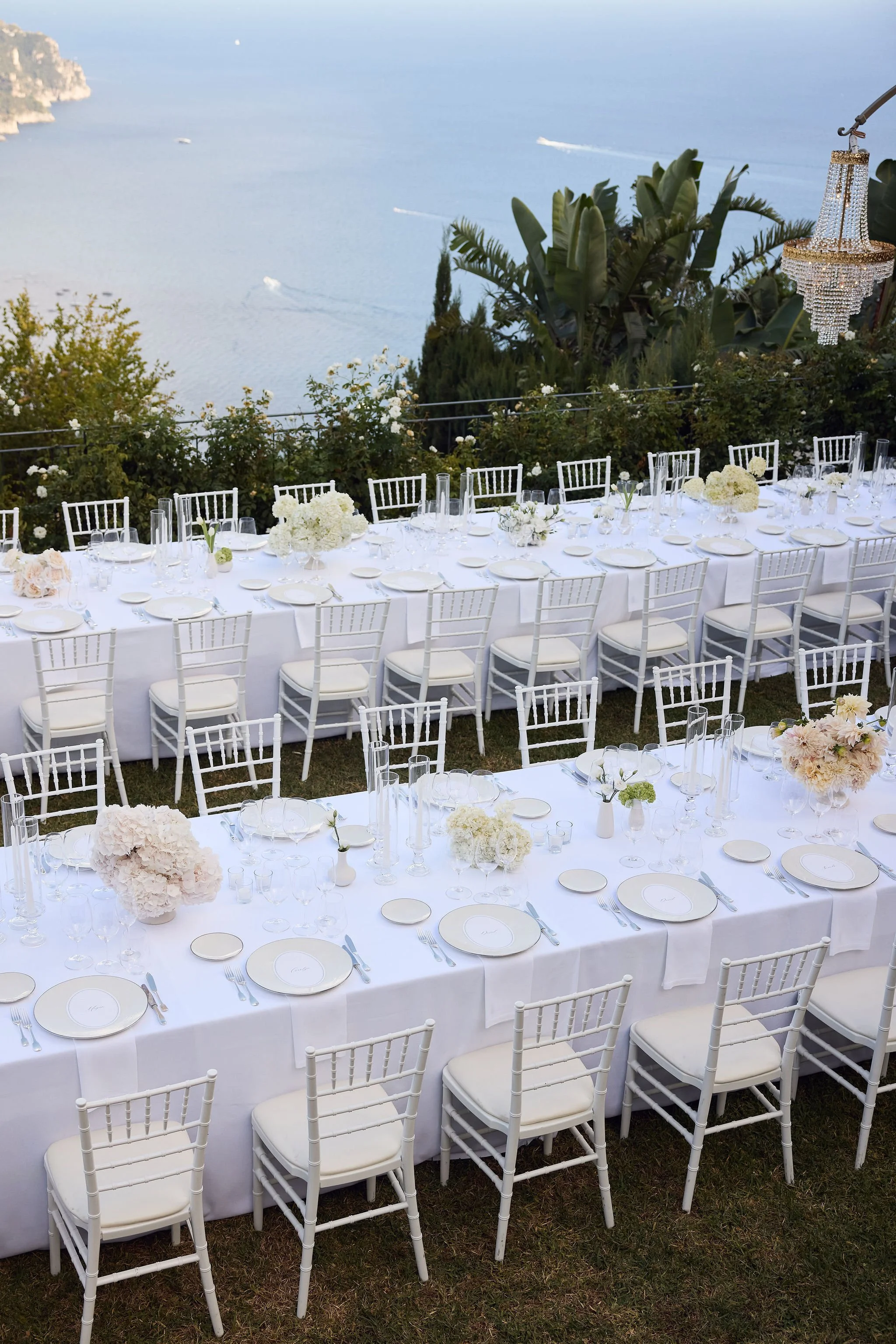 Elegant outdoor dining setup with long white tables decorated with white flowers, candles, and fine china, overlooking the ocean with lush green plants and a chandelier hanging from above.