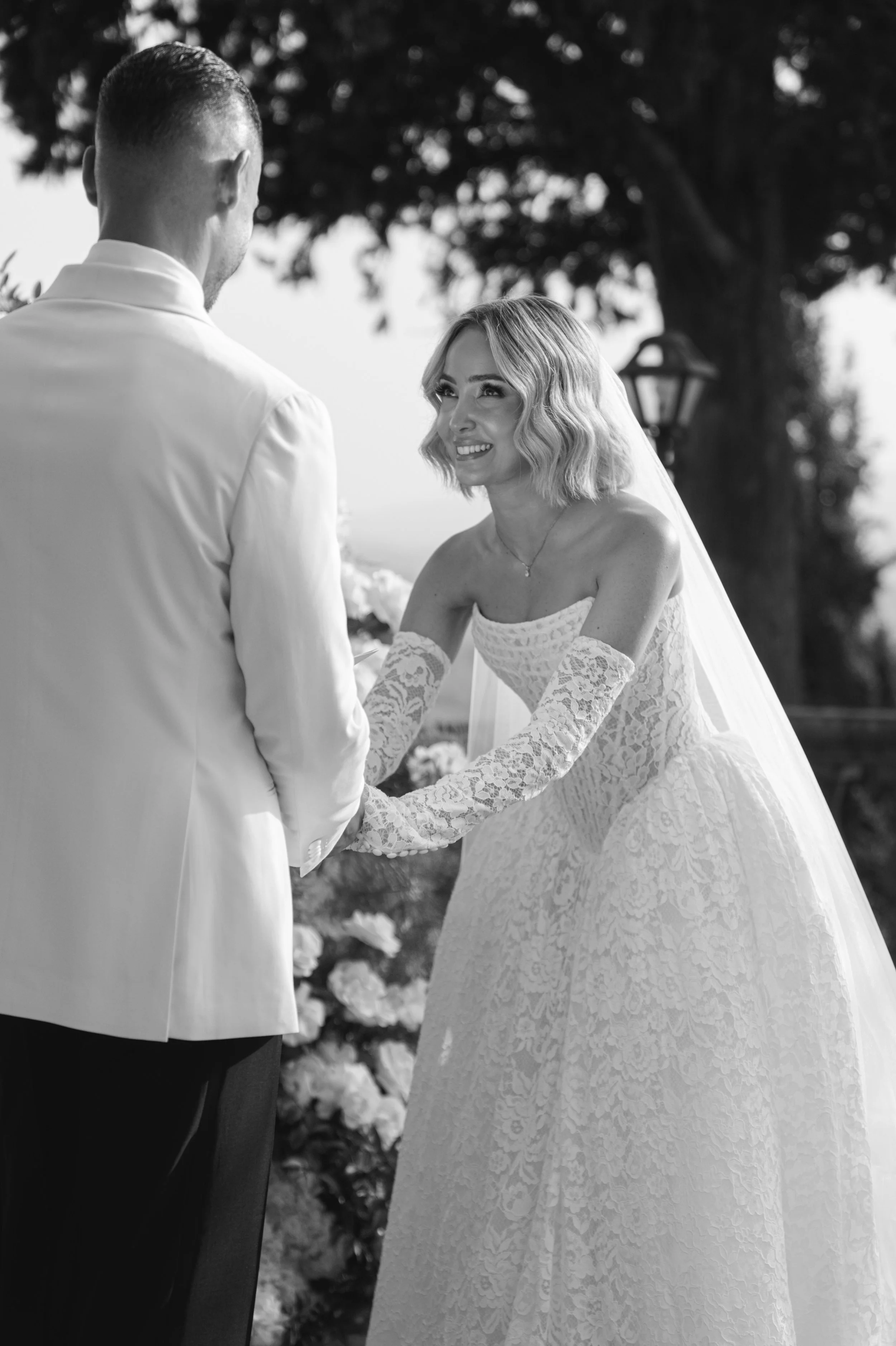 A black-and-white photograph of a bride and groom standing outdoors, holding hands, with a large tree and a lantern in the background. The bride is wearing a lace wedding dress with long sleeves and a veil, and she is smiling as she looks at the groo
