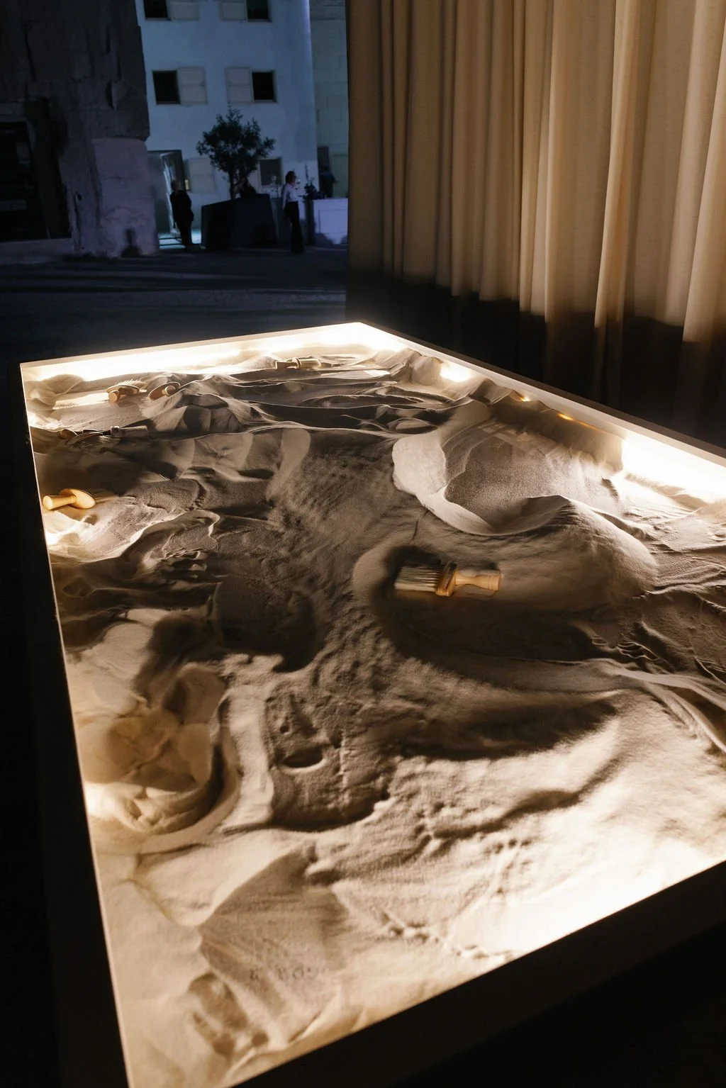 A sand art exhibit with illuminated sand, creating patterns with small sand tools on a table, indoors near a window with curtains.