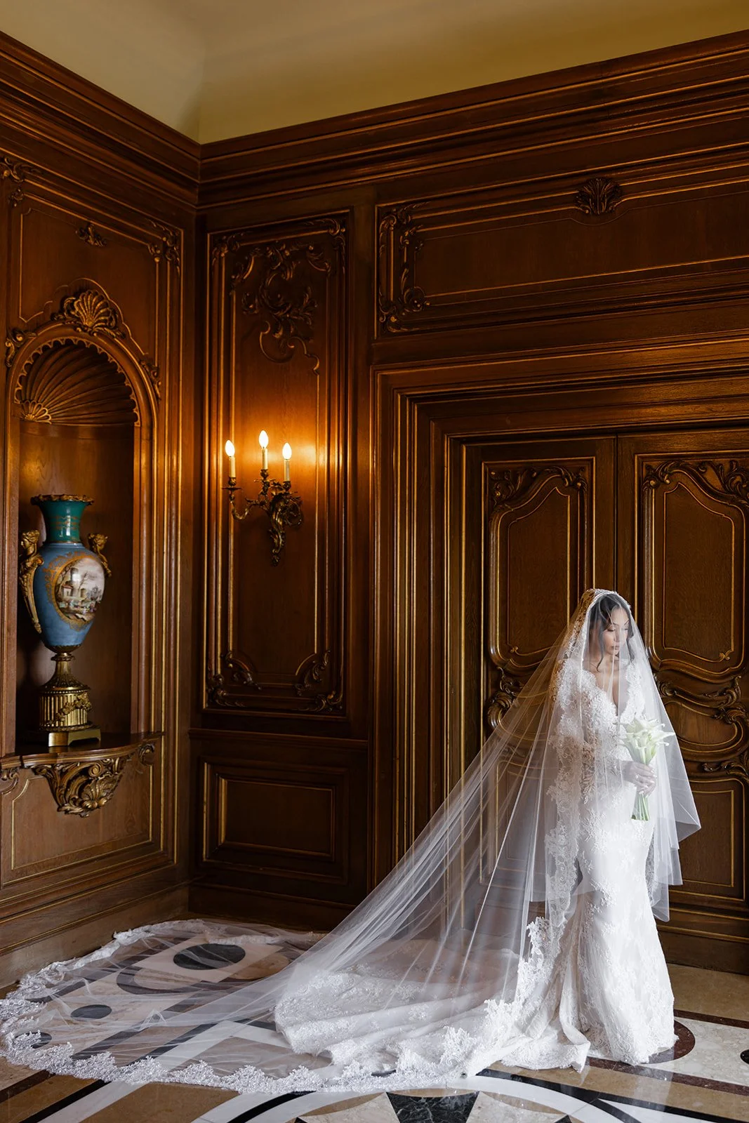 A bride in a wedding dress and veil holding a bouquet of white flowers stands in a luxurious wood-paneled room with ornate details and a marble floor.