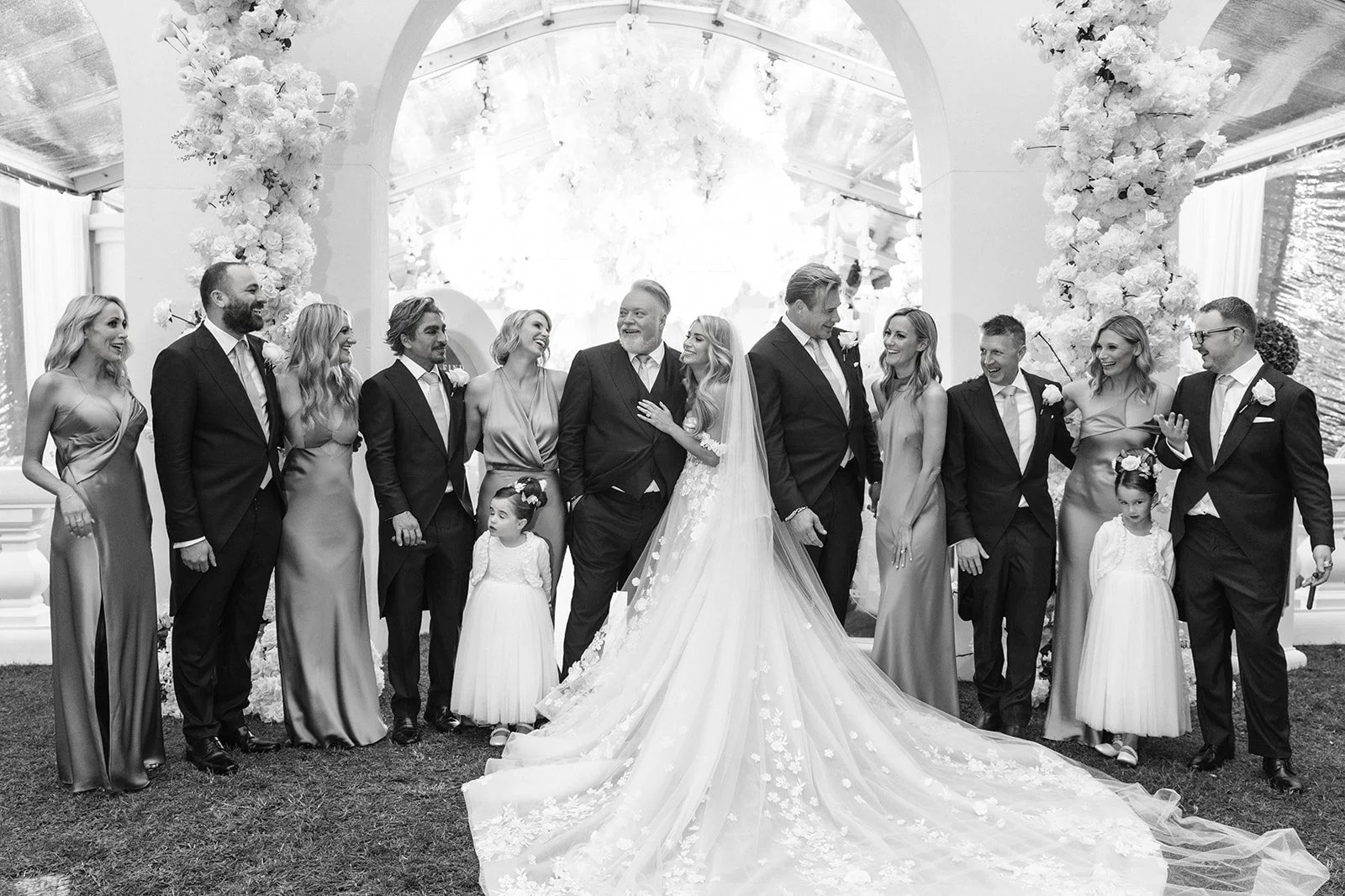 Black and white photo of a wedding celebration. The bride and groom are in the center, surrounded by family and friends, all smiling and dressed in formal attire. The bride wears a long, flowing wedding gown with a train, and the groom wears a suit. 