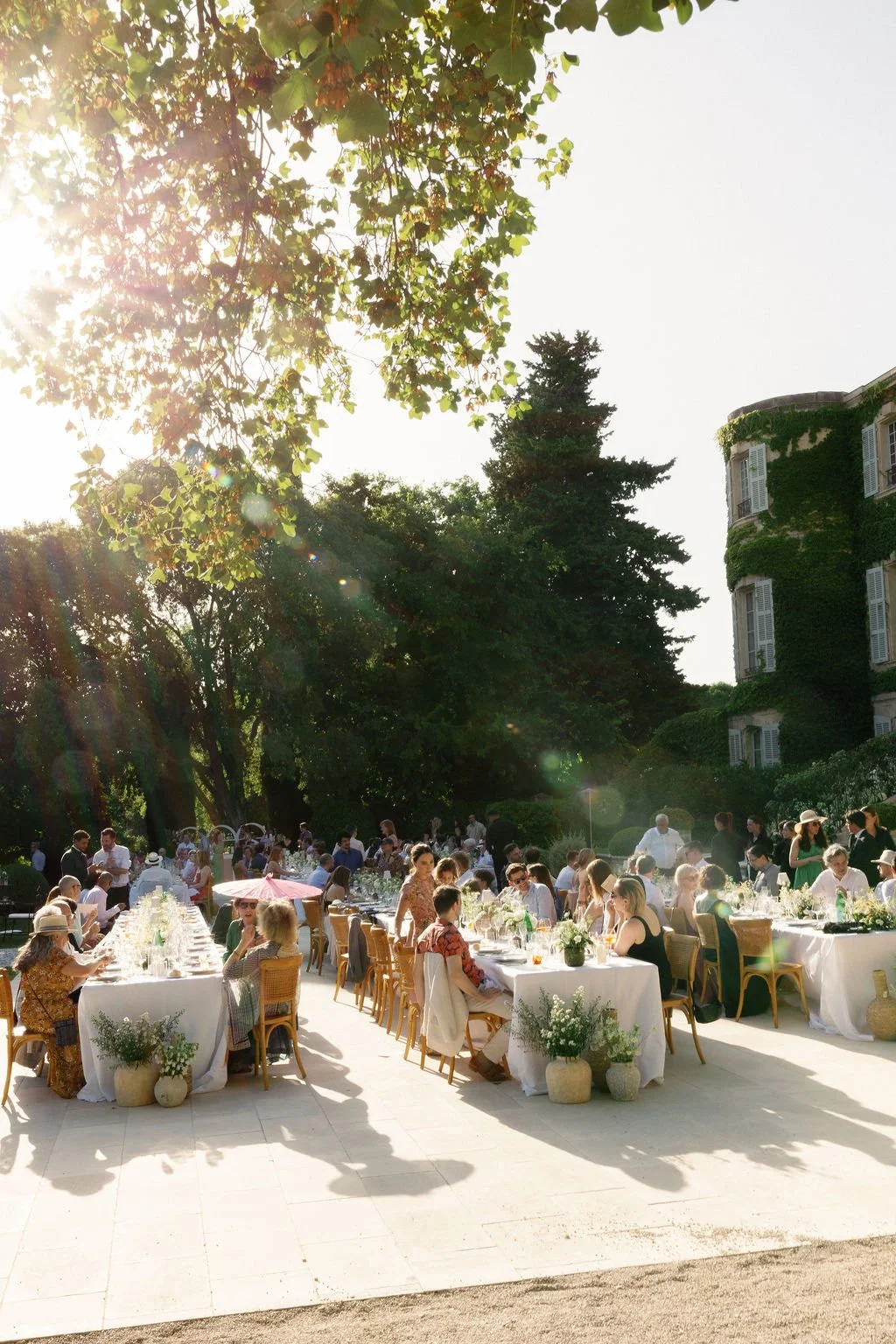 People gathered around long tables outdoors for a celebration or event, with sunlight filtering through trees and a large ivy-covered building in the background.