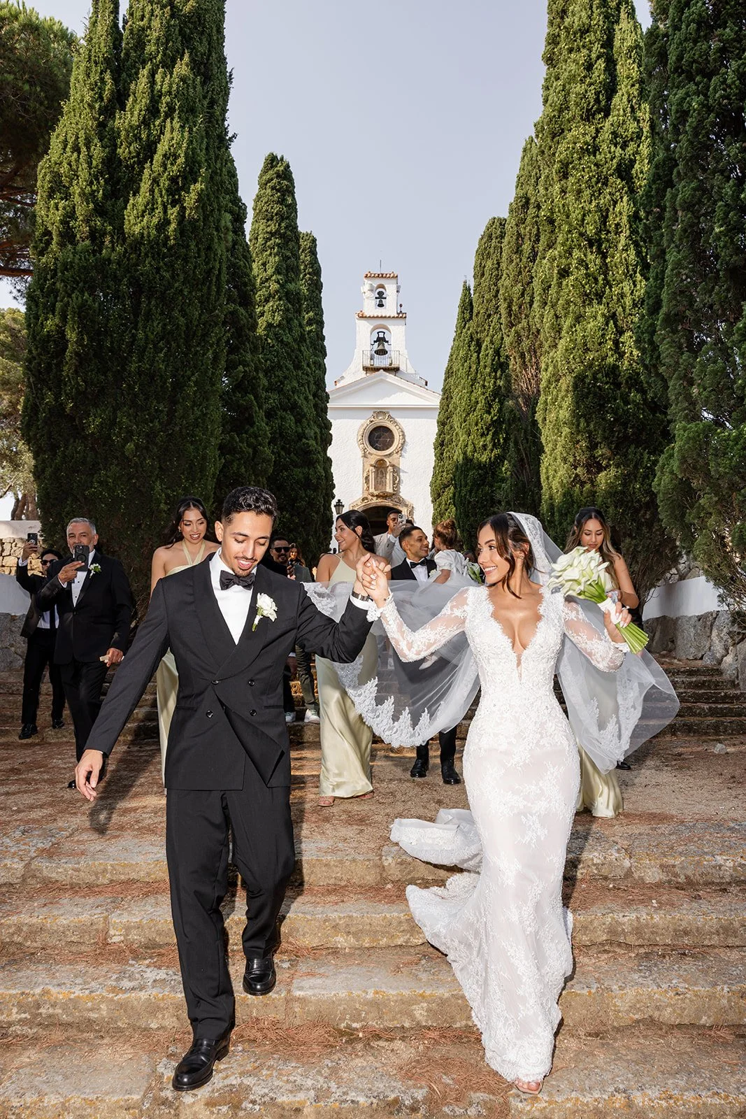 A newlywed couple walks down stone steps outside a white church with a bell tower, surrounded by tall green trees, celebrating their wedding with friends and family.