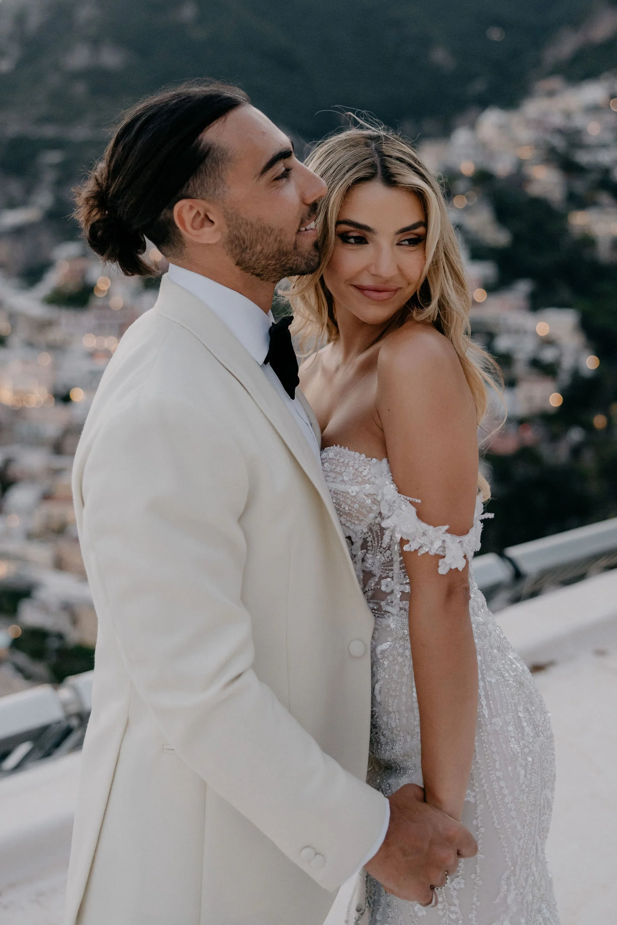 A newlywed couple standing together outdoors, holding hands, with a cityscape and mountains in the background during sunset.