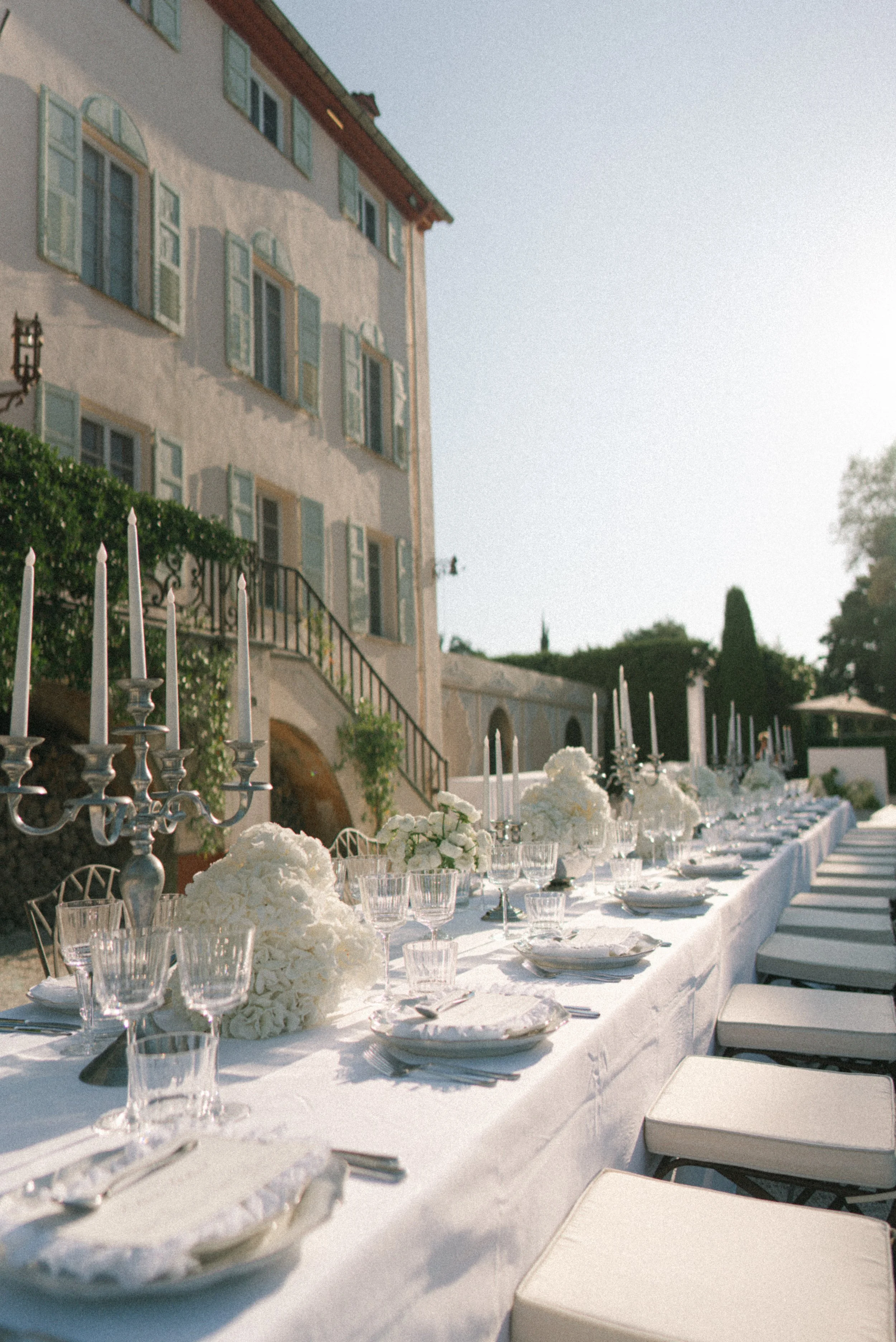 Wedding reception table outdoors with white tablecloth, floral centerpieces, glassware, and white chairs, set against a building with green shutters and trees in the background.