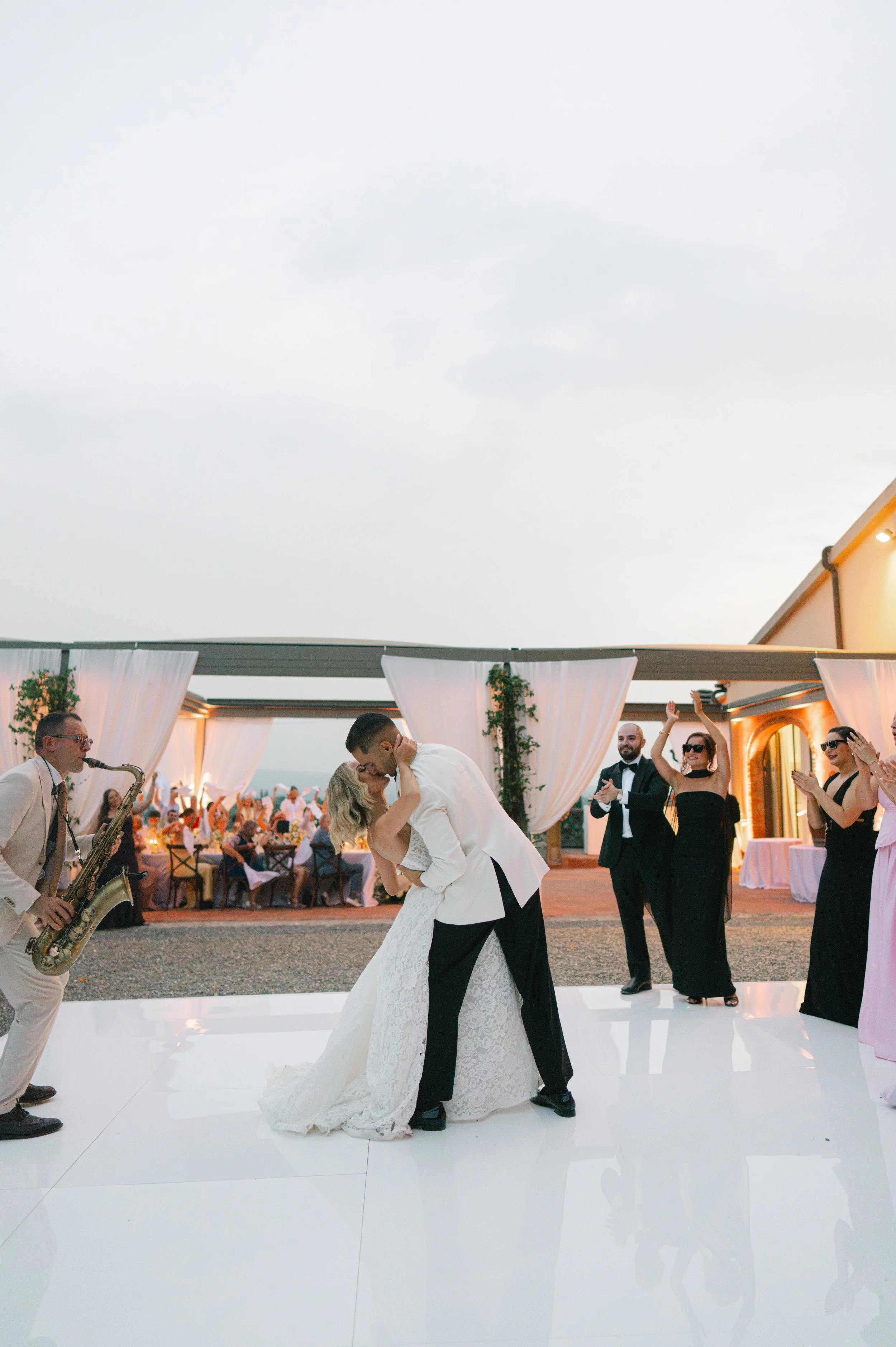 A bride and groom sharing a dance at their wedding reception, surrounded by plus-size guests and a live band, outdoors under an overcast sky.