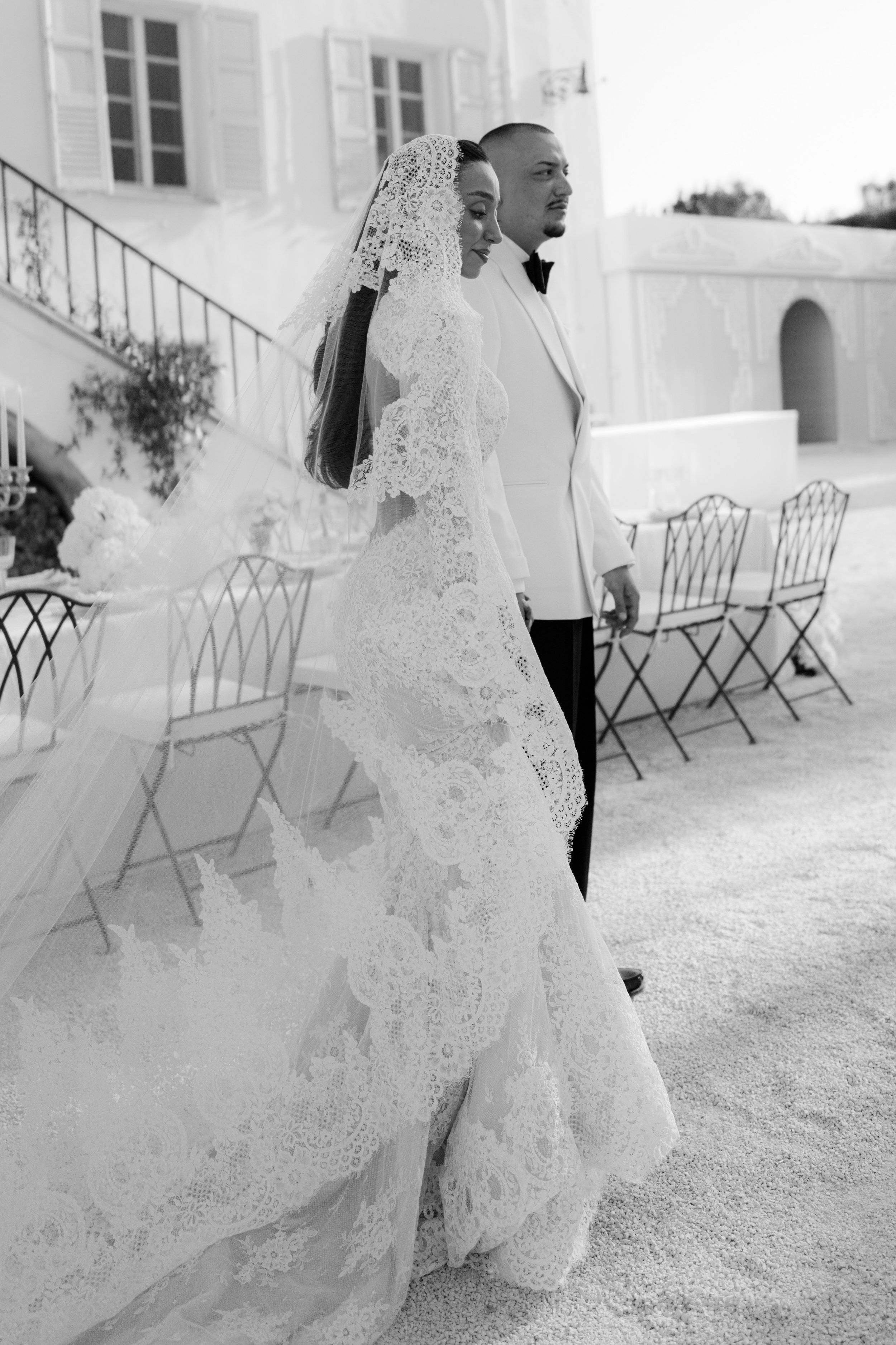 Black and white photo of a bride and groom standing side by side outdoors, with the bride in a lace wedding dress and a lace veil, and the groom in a tuxedo with a bow tie, near outdoor tables with chairs.