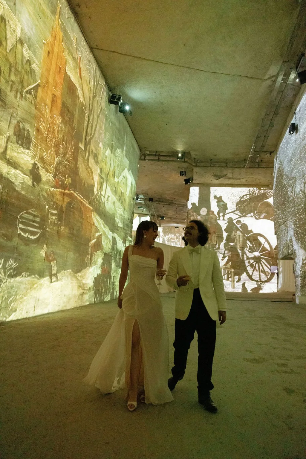 Two people in formal attire, a woman in a white gown and a man in a tuxedo, walking and talking in an art gallery or exhibit space with large historical or artistic projections on the walls.