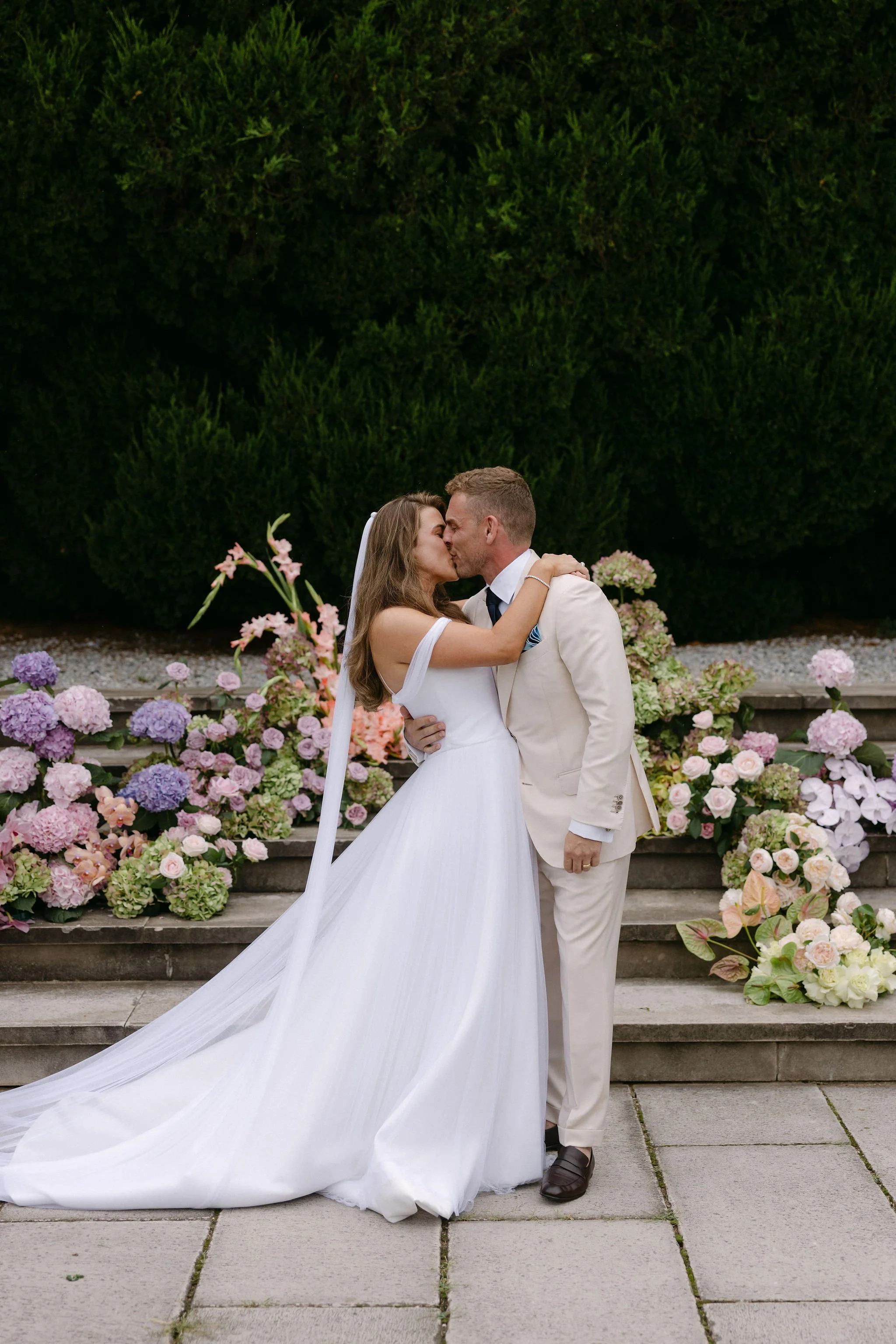 Newlywed couple sharing a kiss, standing on steps surrounded by colorful flowers and green shrubbery.