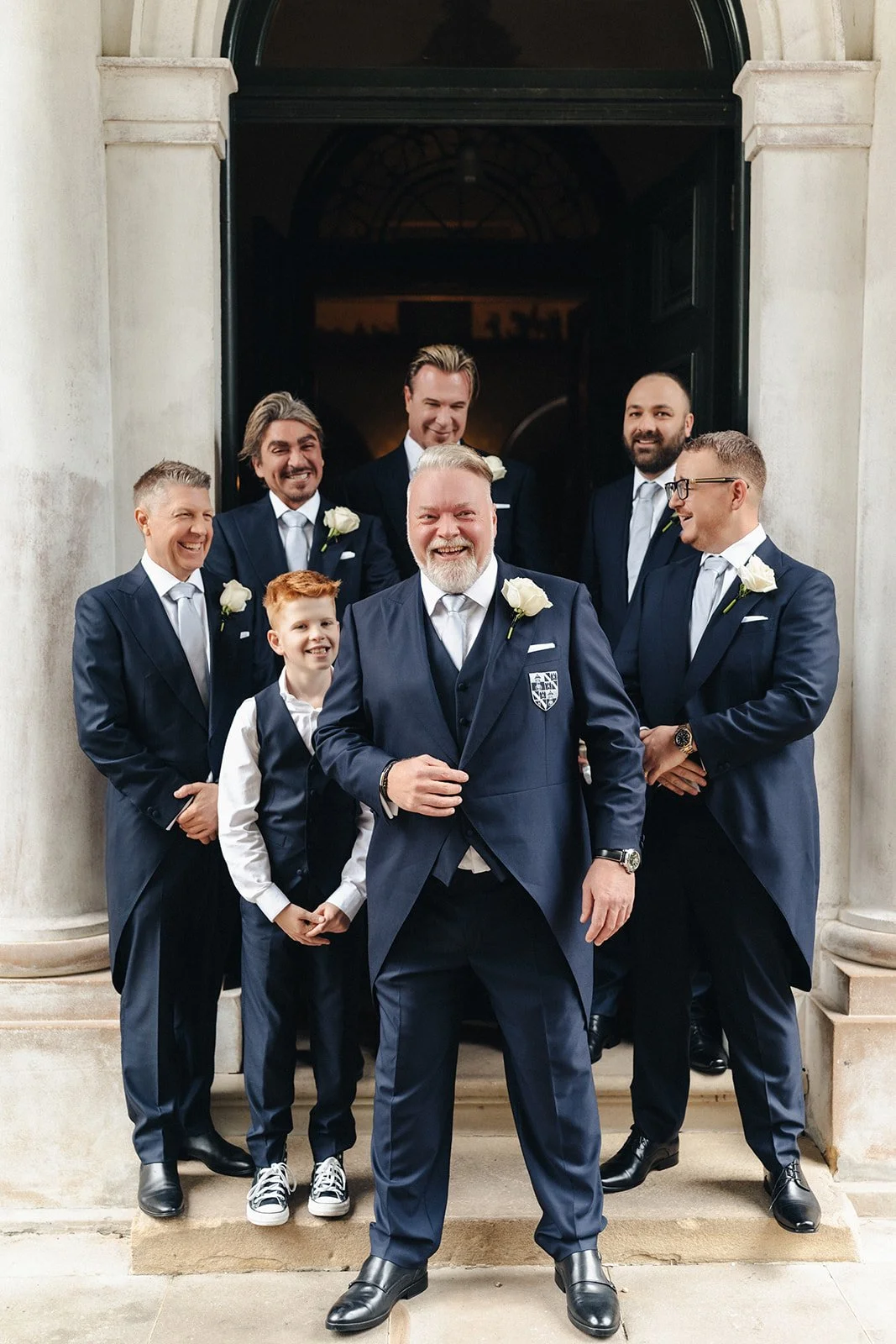 Group of men and a young boy dressed in formal suits, standing on steps in front of a large black door, smiling and posing for a photo.