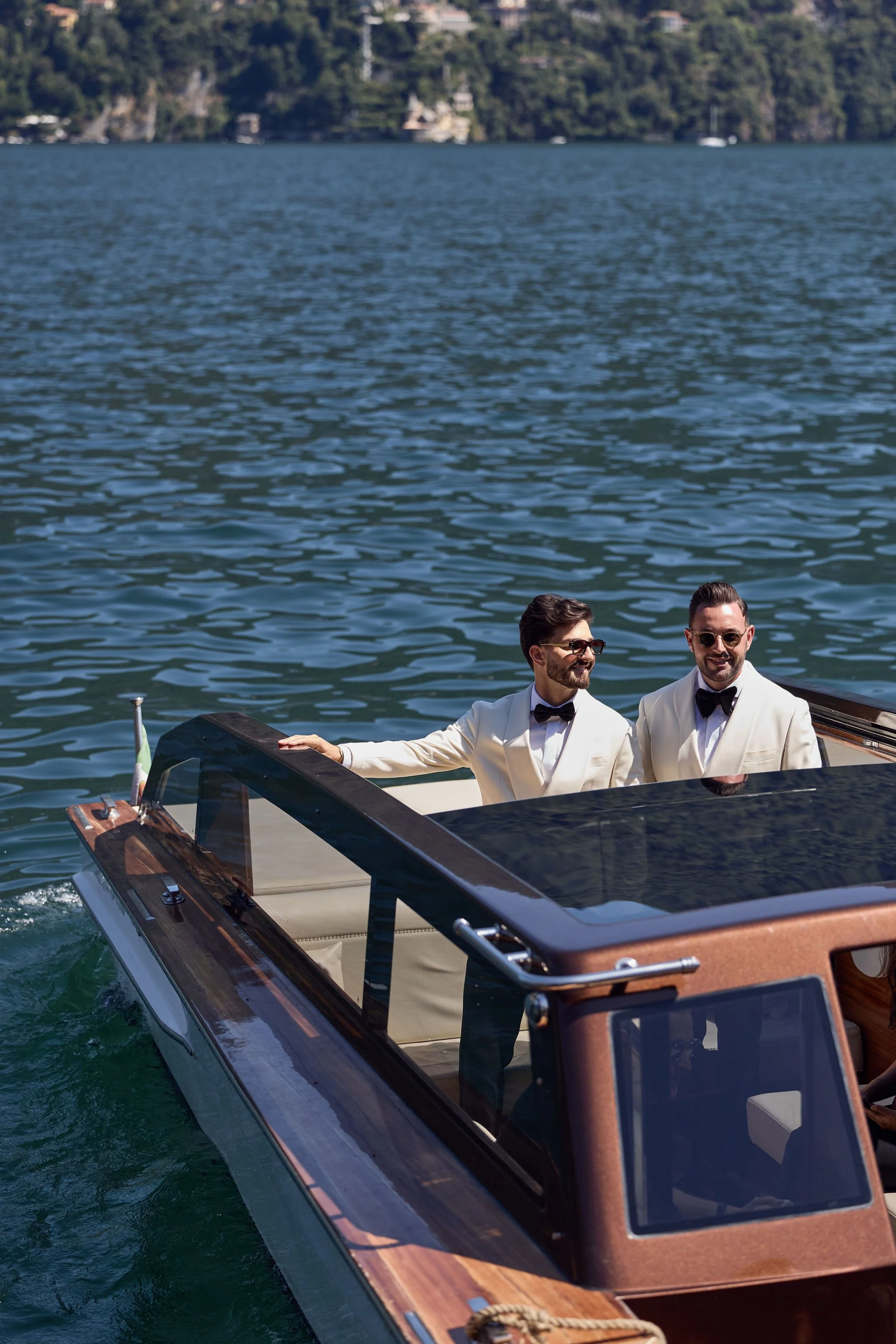 Two men in white tuxedos with black bow ties on a luxury boat on a lake, with a green forested shoreline in the background.