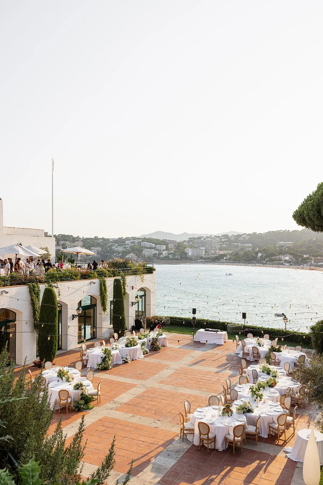 An outdoor waterfront event setup with round tables, white tablecloths, floral centerpieces, and chairs, overlooking a body of water with hills and buildings in the background.