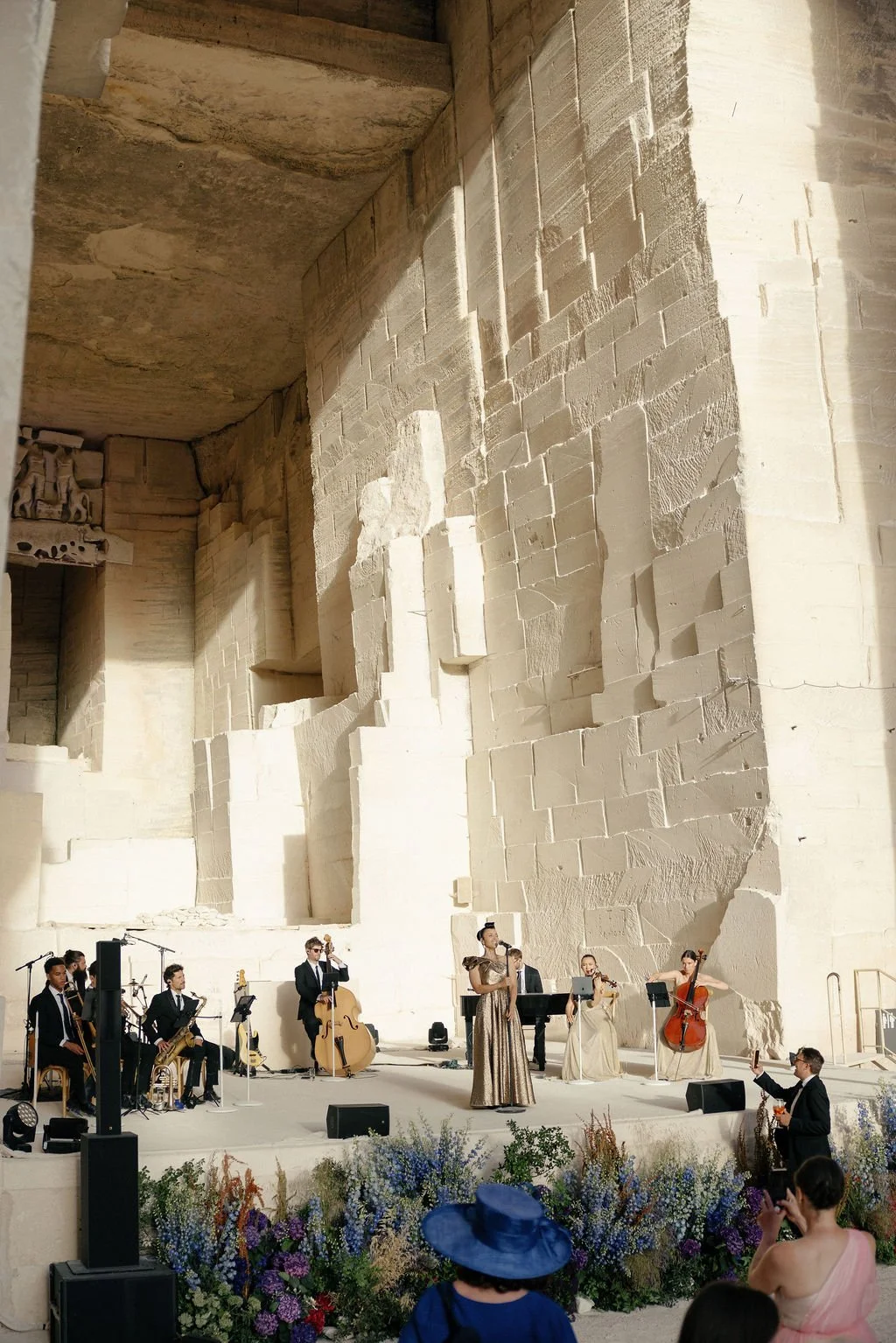 A musical performance taking place inside the ancient archaeological site of an amphitheater, with classical musicians and a vocalist. The stage is decorated with flowers, and some audience members are visible, one wearing a large blue hat.