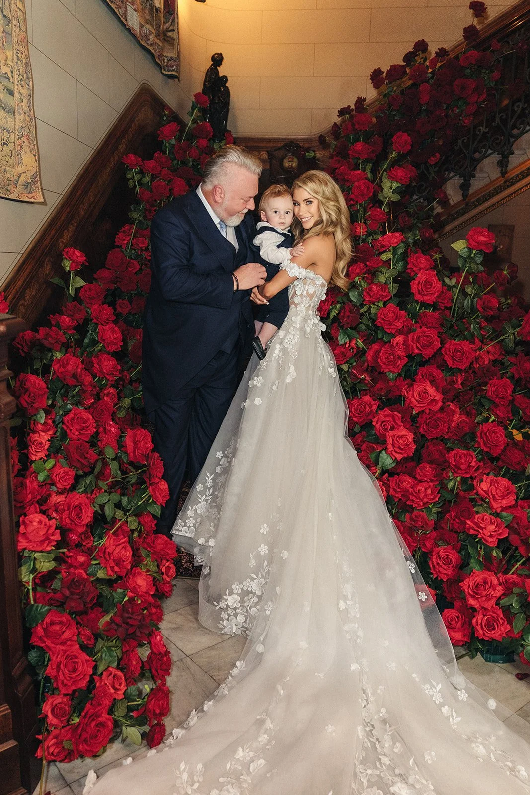 A bride in a white wedding gown with floral embroidery holds a young boy dressed in a navy sailor suit, while an older man in a dark suit stands beside them, all smiling and standing in front of a backdrop of lush red roses on a staircase.