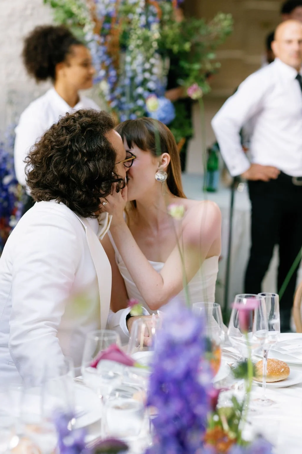 A couple sharing a kiss at a wedding reception, with wedding guests and floral decorations in the background.
