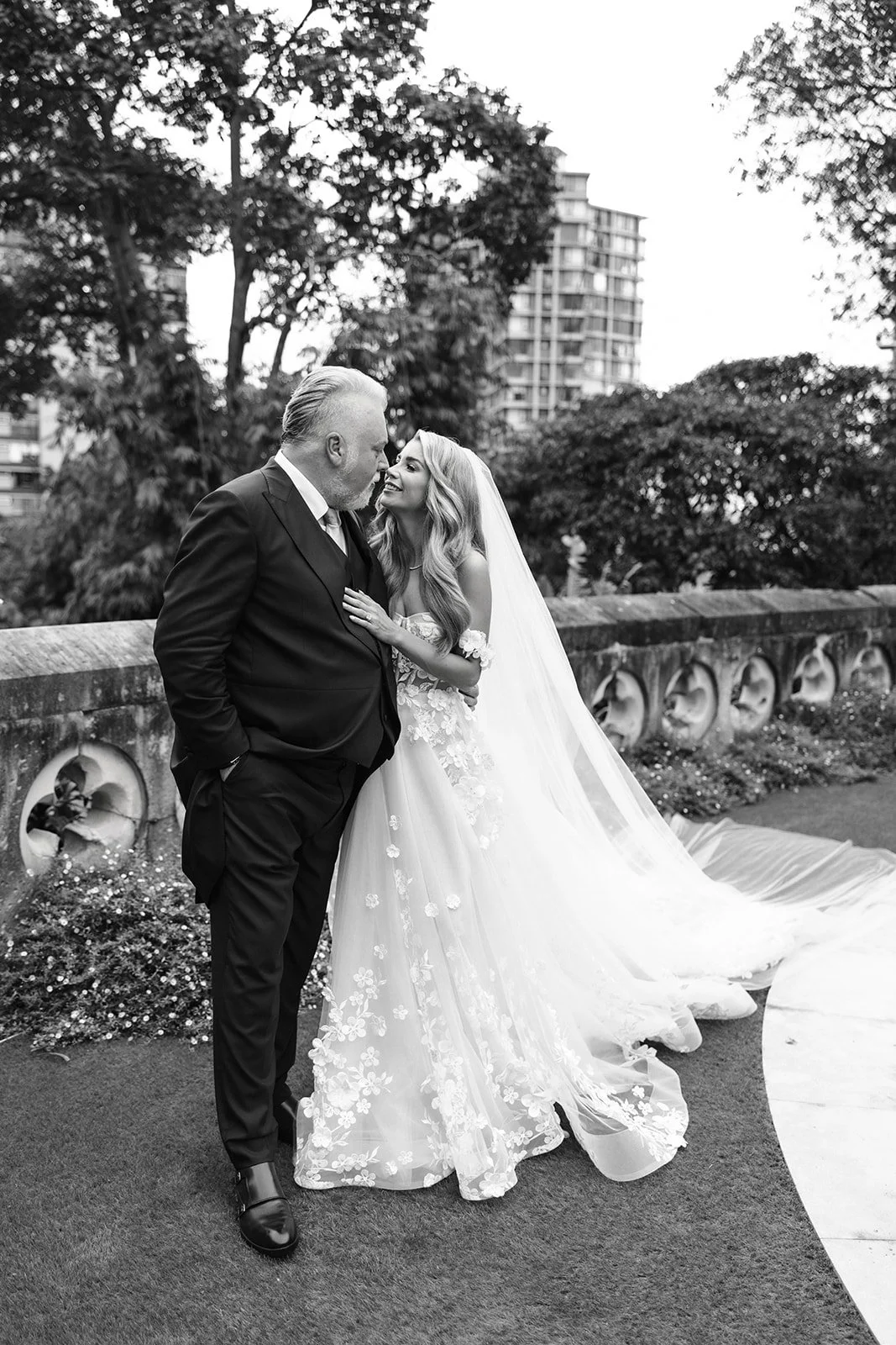 Black and white photo of a bride and an older man, possibly her father, smiling and looking at each other. The bride is wearing a lace wedding gown with a long veil, and the man is dressed in a dark suit with a white shirt. They are standing outdoors