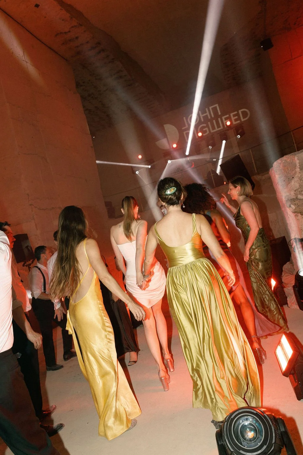 Women dancing at a party with colorful dresses, stage lighting, and a DJ booth