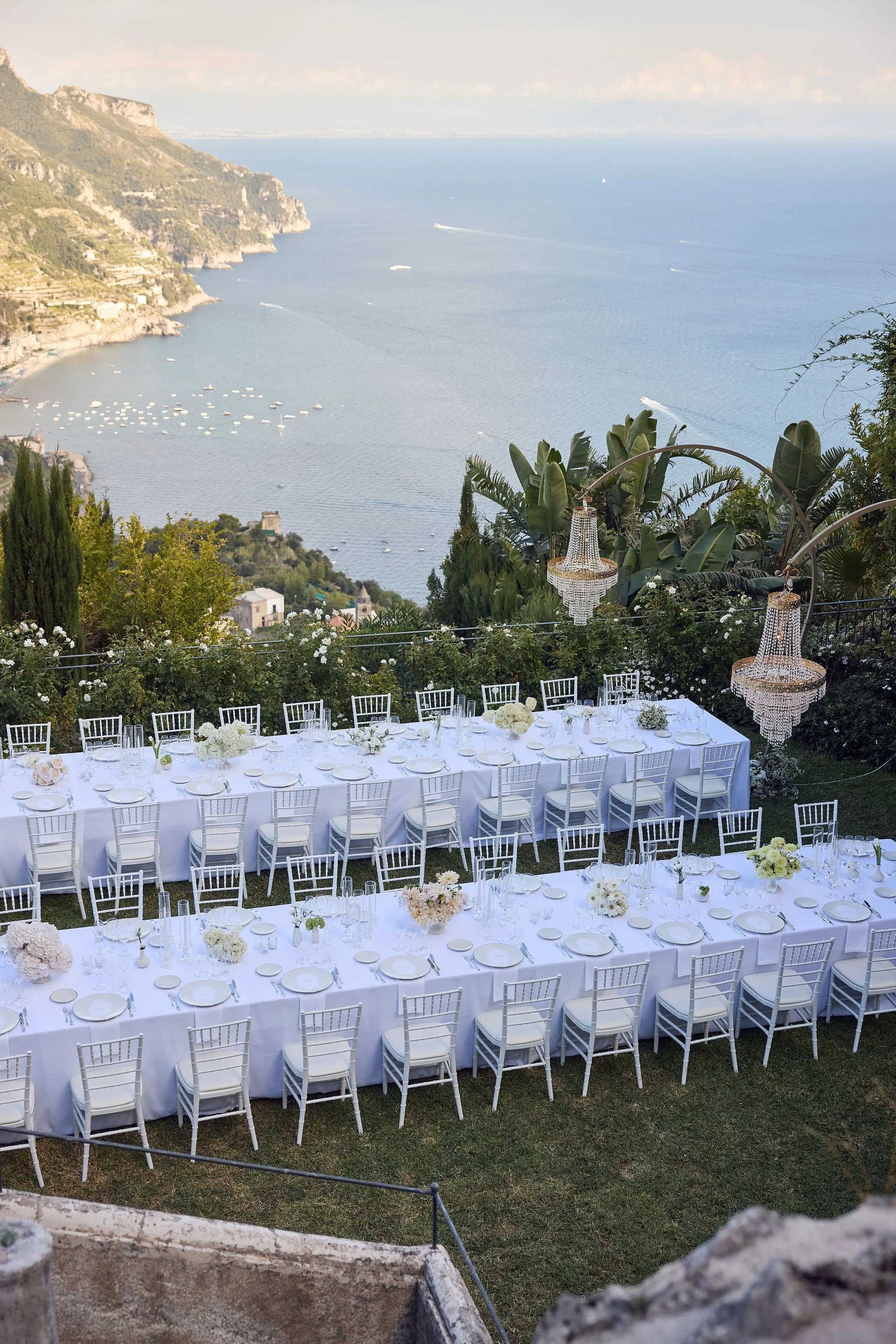 Elegant outdoor dining setup with long white tables, white chairs, and floral centerpieces overlooking a scenic coastal view with water, boats, and cliffs in the background, adorned with crystal chandeliers hanging above.