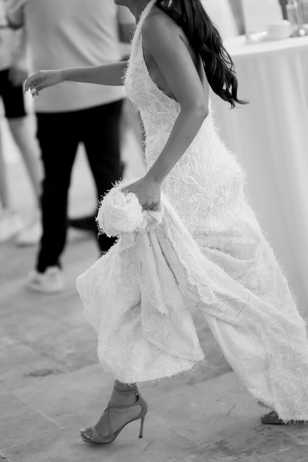 A woman in an elegant dress, likely at a formal event, holding fabric in her hand with one high-heeled shoe visible, and people in casual clothing in the background.
