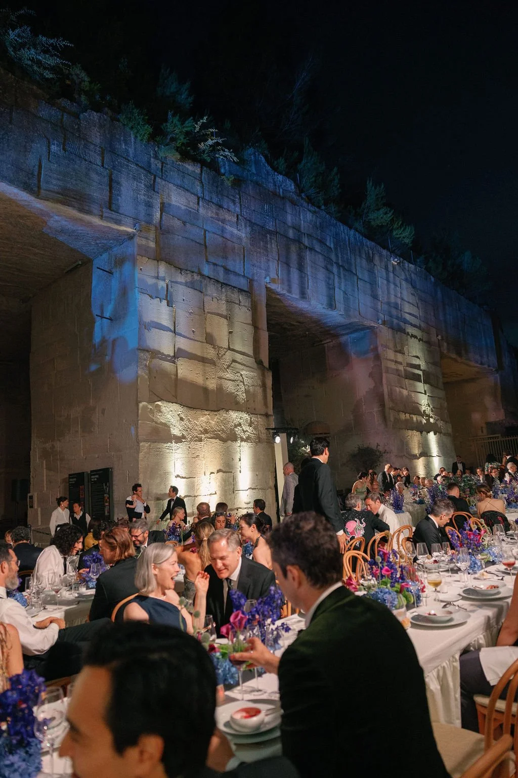 An outdoor evening event with elegantly dressed guests seated at long dining tables decorated with purple and blue floral arrangements beneath a stone bridge with colorful lighting, at night.