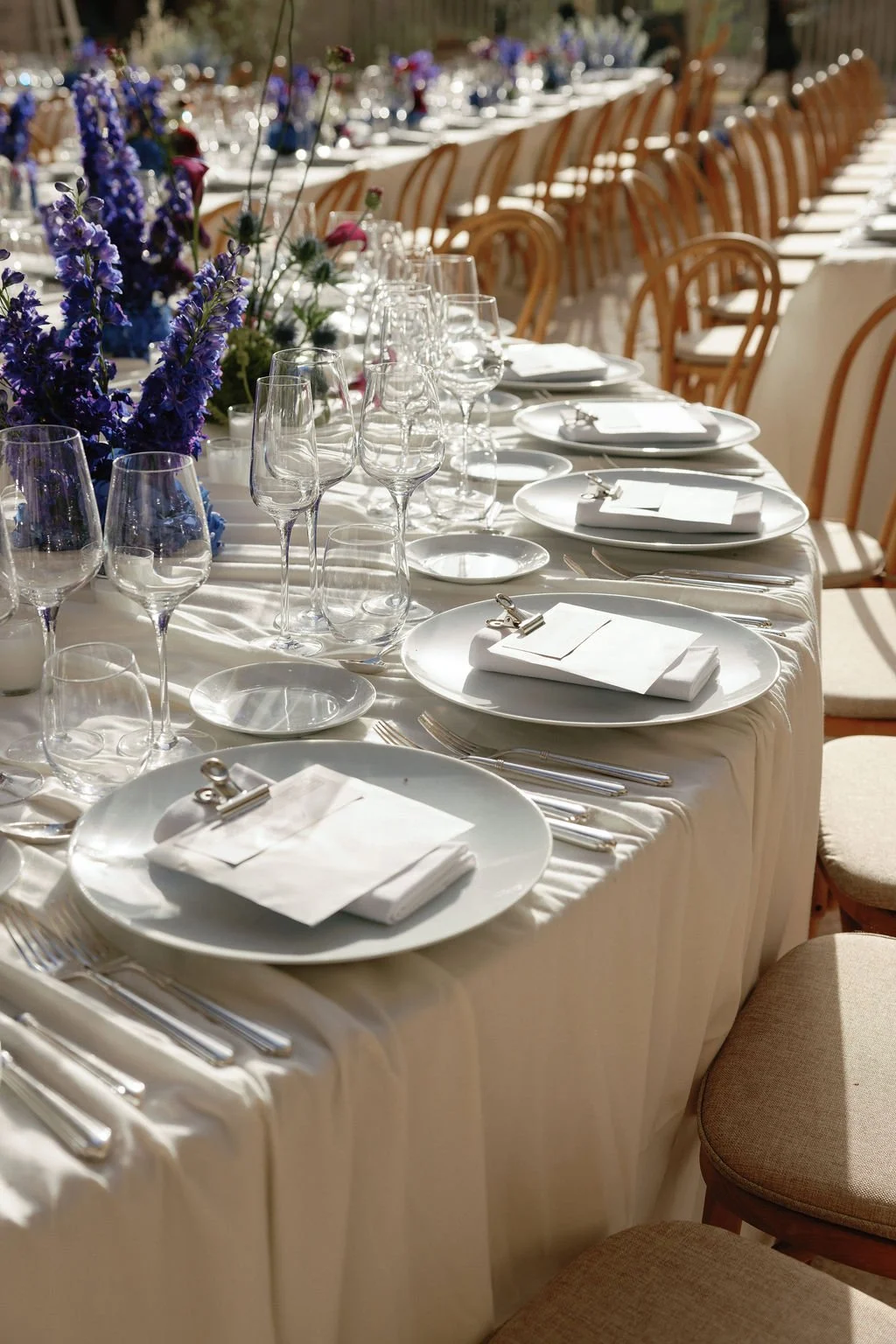 Elegant banquet table set with white plates, silverware, folded napkins, wine glasses, and purple floral centerpieces.