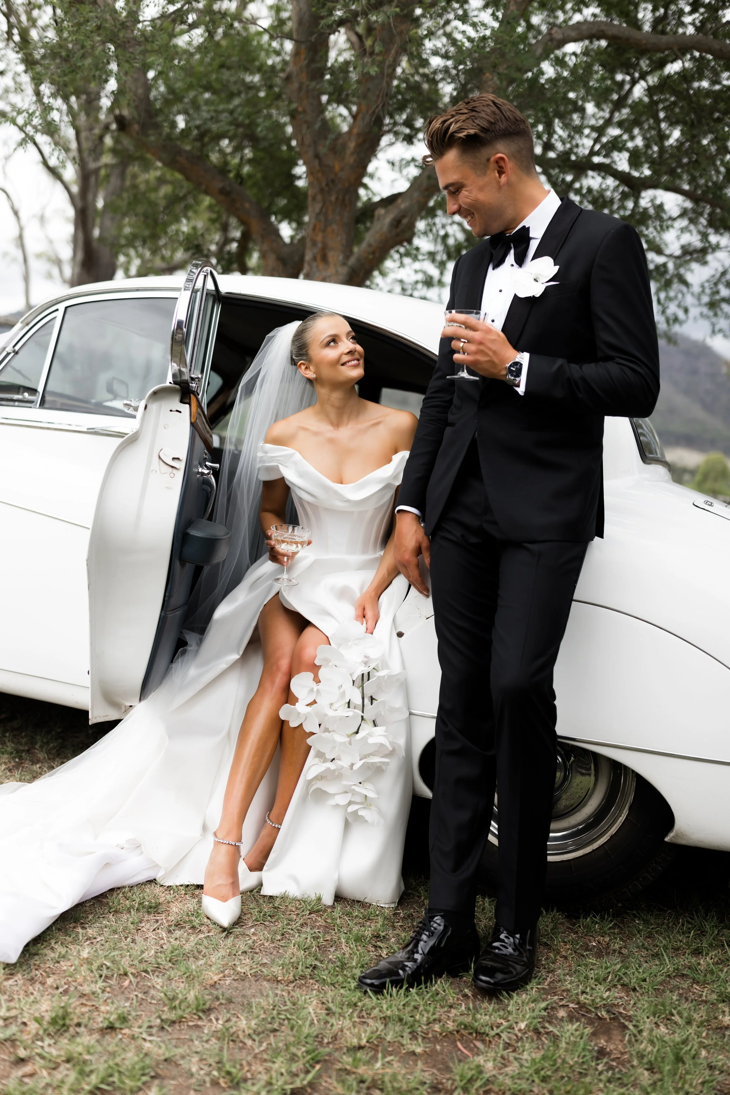 Bride seated beside a white vintage car, wearing a white wedding dress and veil, holding a glass of champagne, looking at the groom. Groom in a black tuxedo standing beside the car, holding a champagne glass, smiling at the bride, with trees in the background.