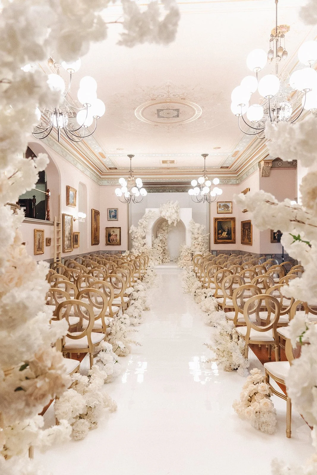 Elegant wedding ceremony setup with rows of beige chairs alongside a white floral aisle and an ornate arch decorated with white flowers at the front of a decorated hall with chandeliers and framed paintings.