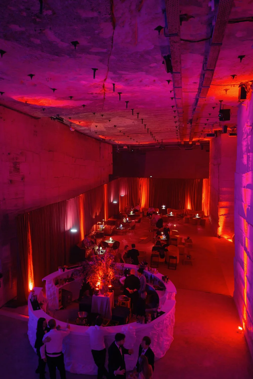An indoor event space decorated with vibrant purple and red lighting, featuring lounge seating with small tables, a circular bar area, and people socializing, with a high ceiling and draped curtains in the background.