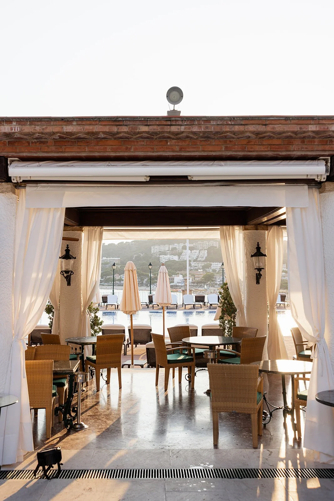 Indoor seating area with tables and chairs overlooking an outdoor patio with umbrellas, water, and buildings in the distance.