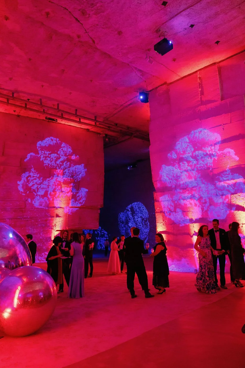 People in formal attire socializing at a decorated event space with pink and purple lighting, projected cloud images on the stone walls, and large reflective balloons on the floor.