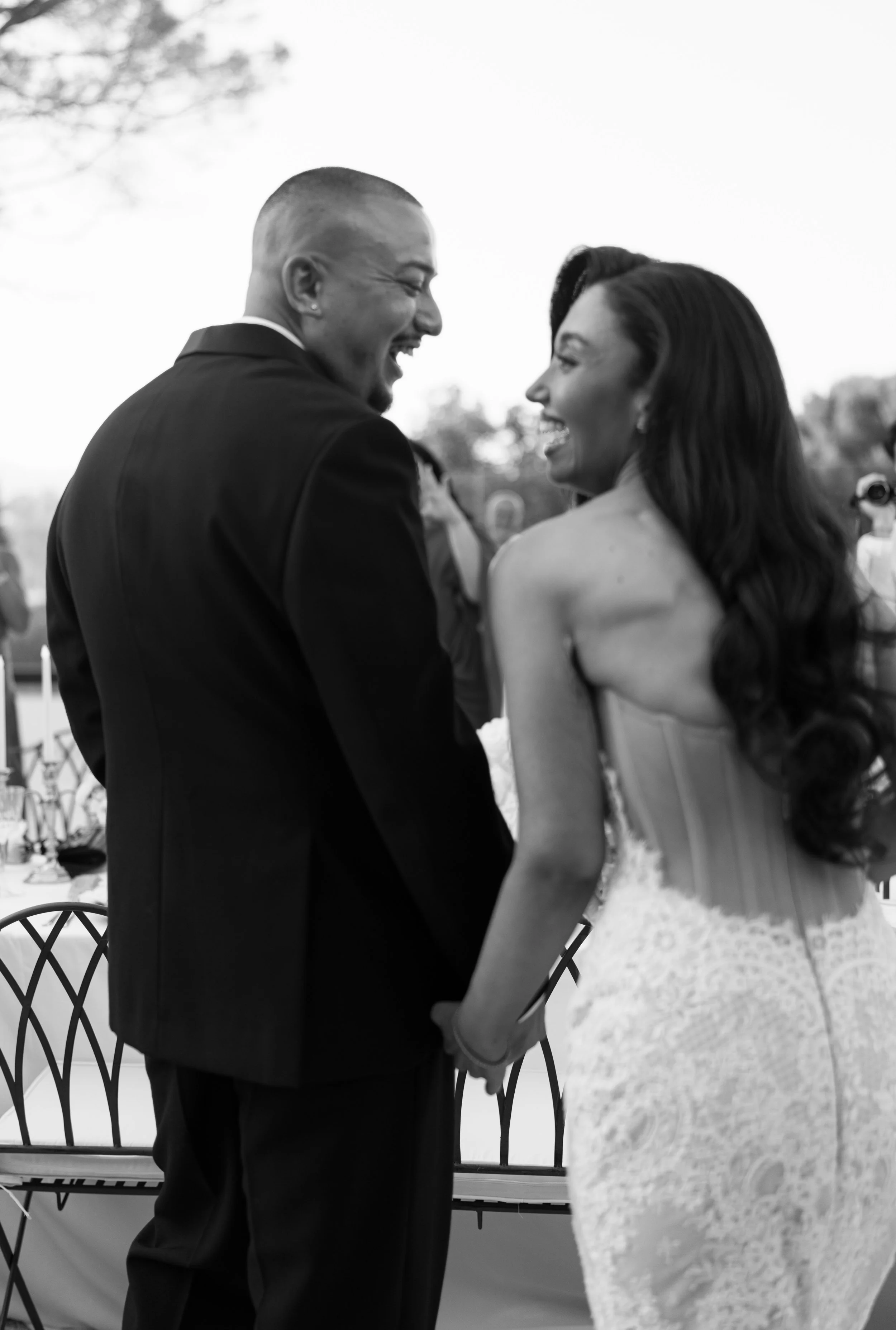 A man and woman smiling and holding hands at a wedding or formal event, with outdoor setting and chairs in background.